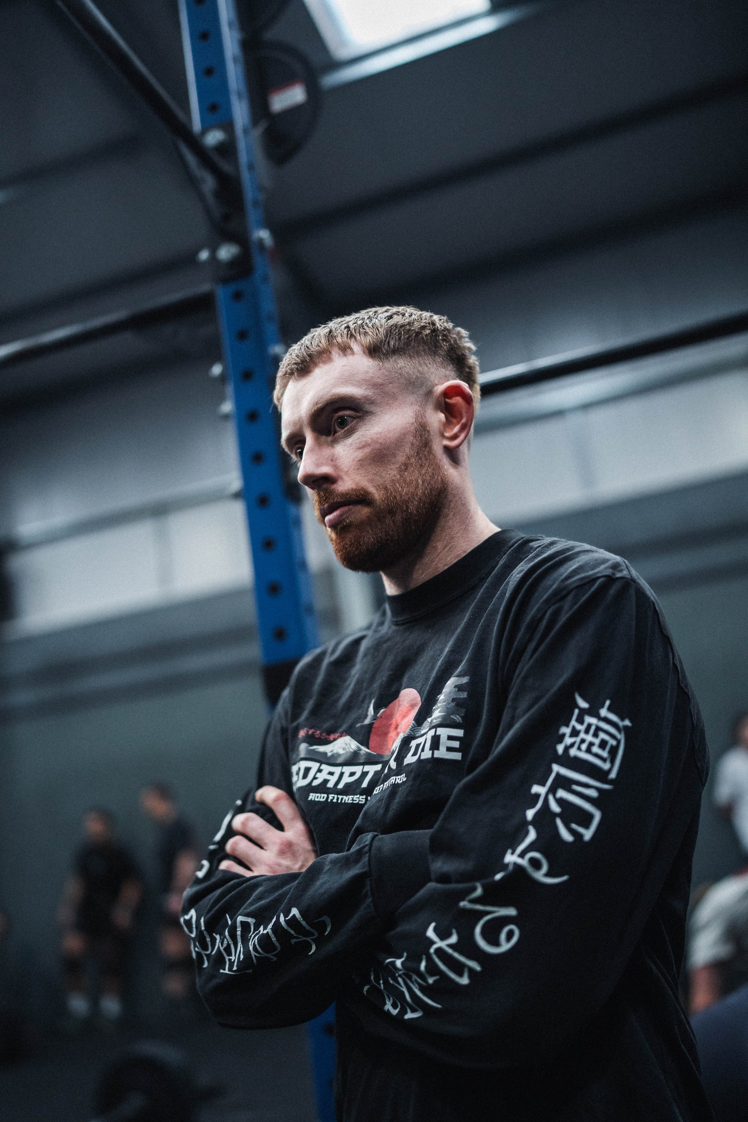 A man with short blonde hair and a beard in a gym, wearing a black hoodie with Asian characters and English text, with arms crossed, standing near gym equipment.