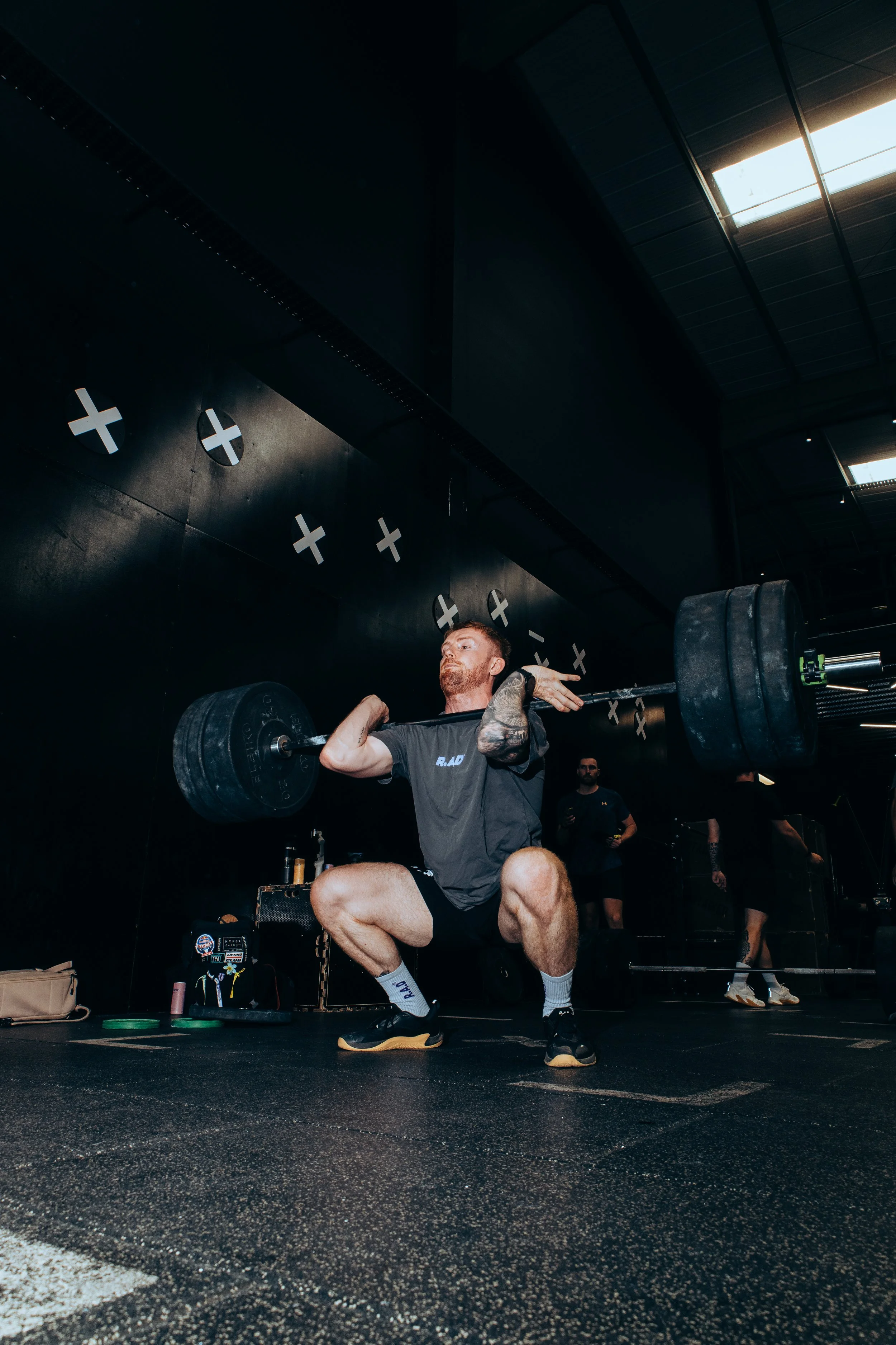 A man with a beard weightlifting at a gym, performing a squat with a barbell loaded with large weights. He is in a black gym with a high ceiling, and others are visible in the background.