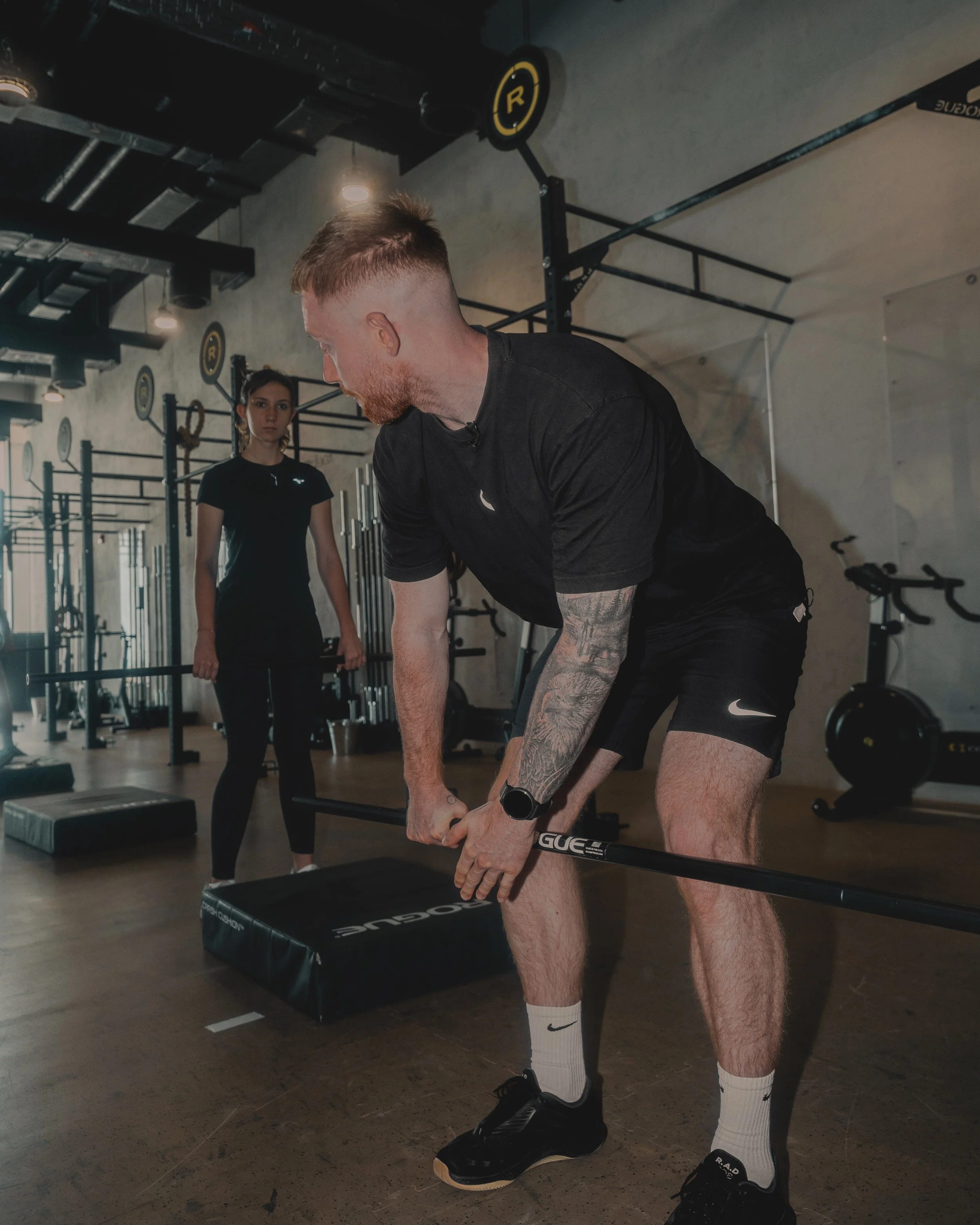 A man with tattoos on his left arm, wearing a black t-shirt, black shorts, and black shoes, is lifting a barbell in a gym. A woman in a black t-shirt and black leggings is standing in the background near gym equipment, observing. The gym has workout stations, a black padded platform, and various equipment mounted on the wall.
