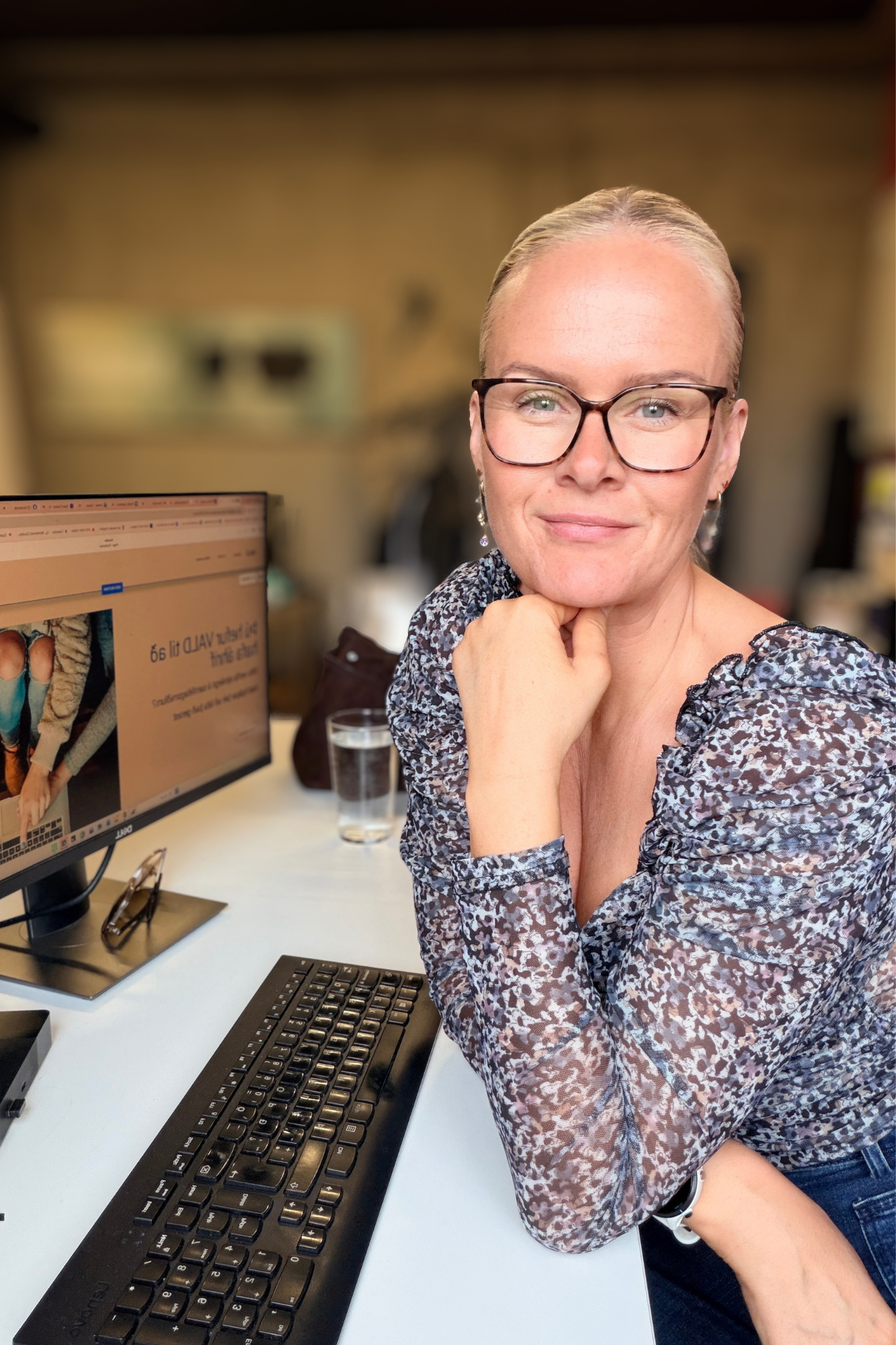 A woman with blonde hair, glasses, and earrings sitting at a desk with a computer monitor and keyboard, smiling at the camera.