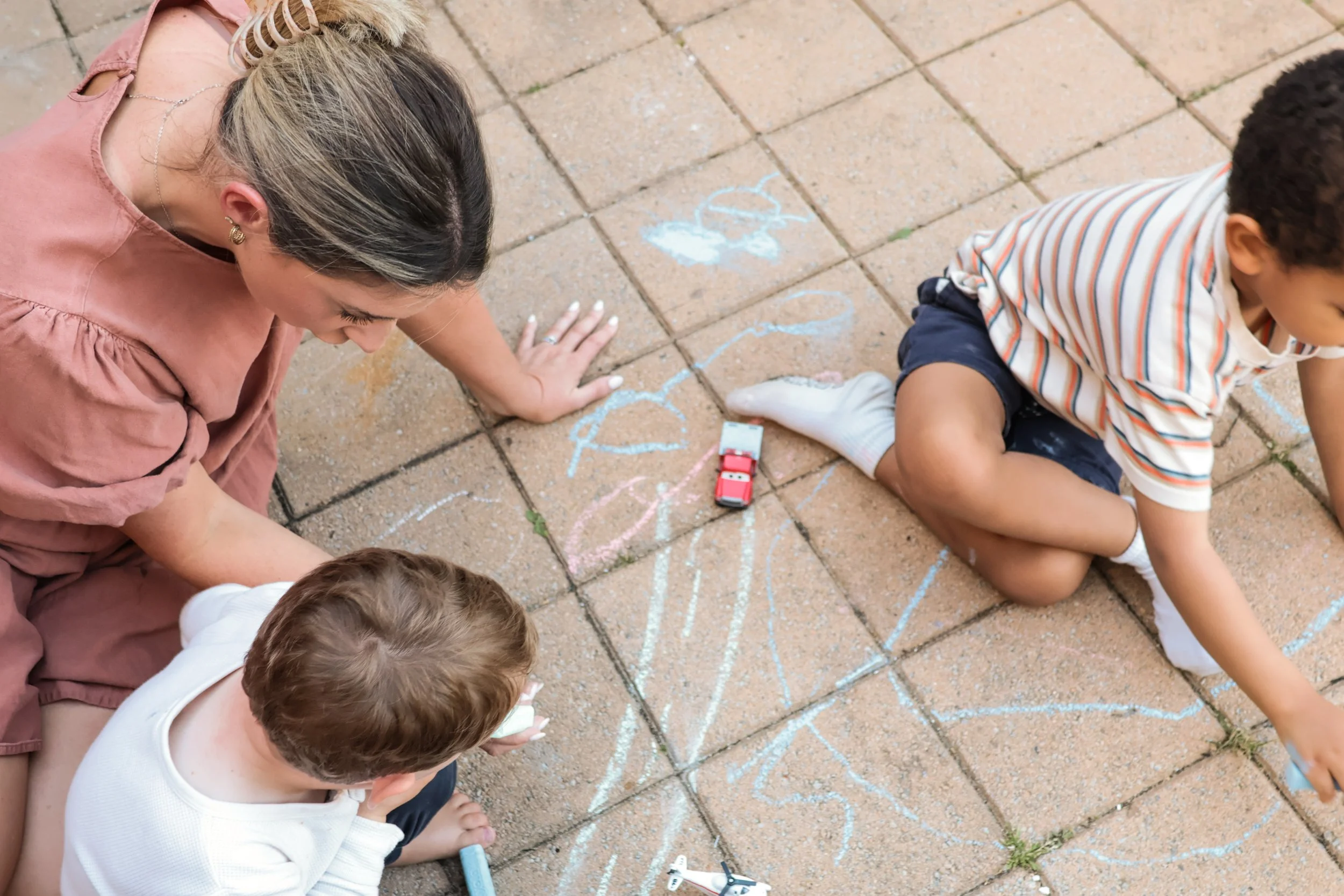 Rachael, with blonde hair and earrings, sitting on the ground, engaging with two young children who are drawing with chalk on a brick patio. The children are sitting and kneeling, focused on their chalk drawings, with toys and chalk scattered around.