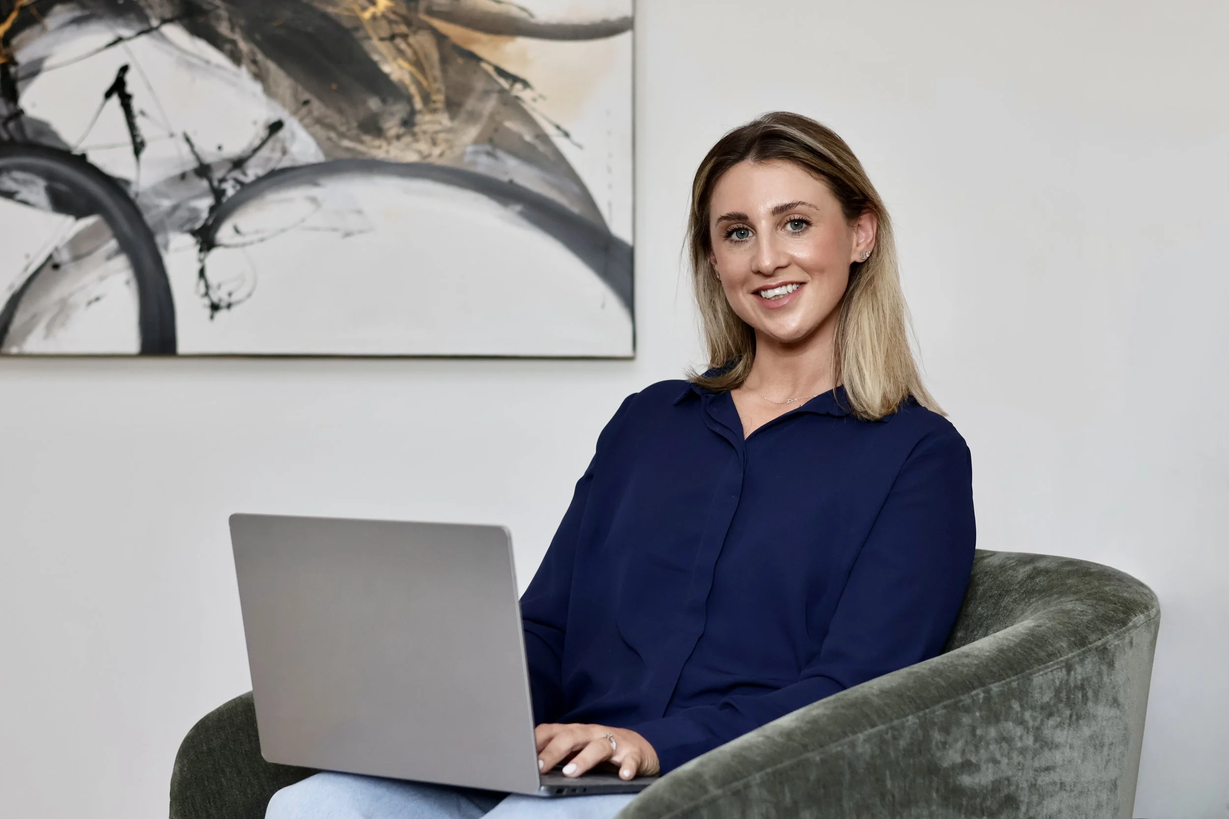 Rachael has dark hair roots, lightening to a golden blonde at the tips. She's wearing a navy shirt and smiling at the camera with a laptop on her knee. She's sitting on a green velvet sofa against a white wall with a framed artwork behind.