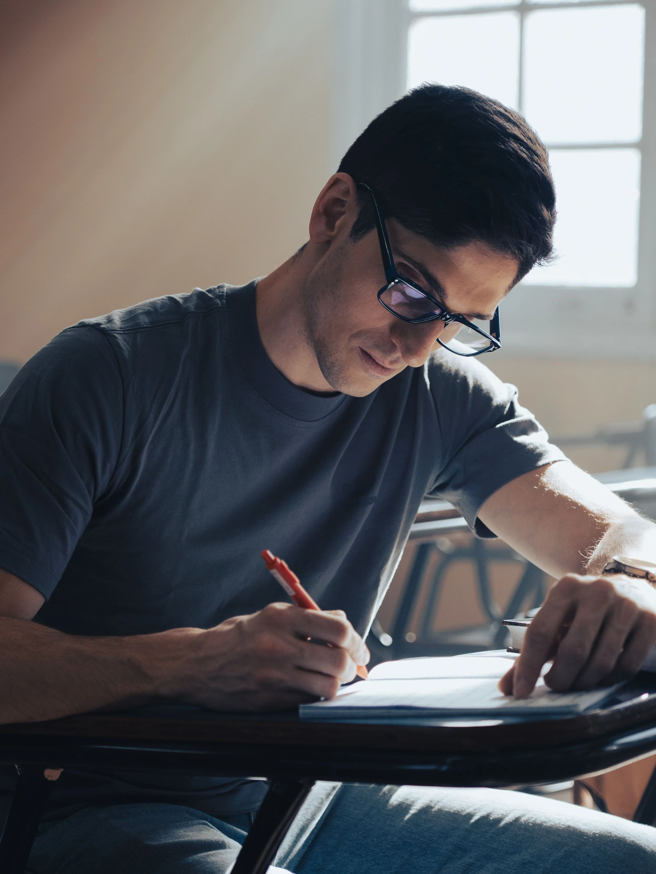 A man wearing glasses sitting at a table, writing in a notebook with a red pen, near a window with natural light.