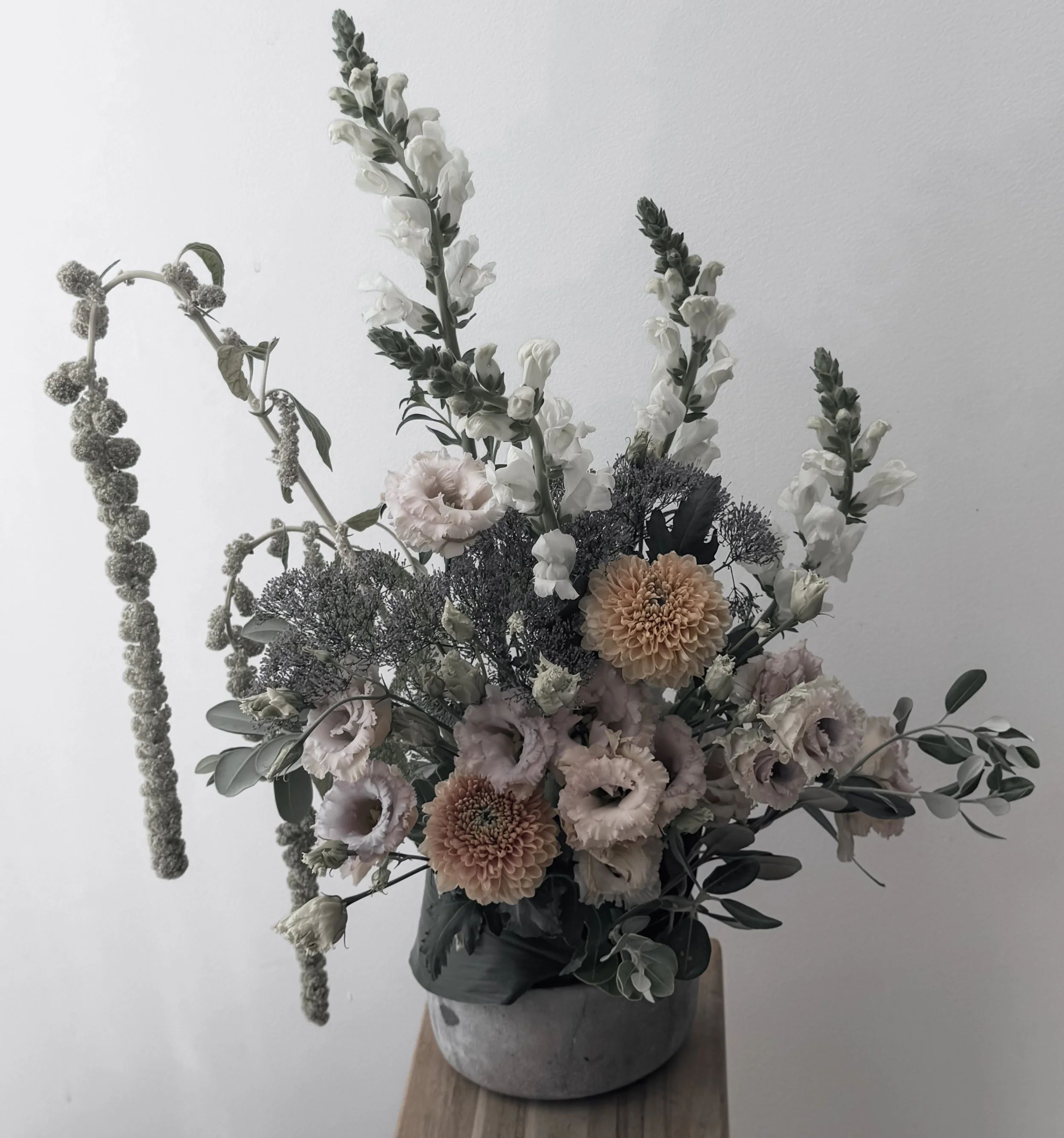 Un arrangement floral dans un vase gris posé sur une table en bois, comprenant des fleurs blanches, des dahlias roses, des feuilles vertes et des éléments gris foncé.