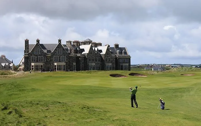 A golfer taking a shot on a golf course with a large, historic-style building in the background.