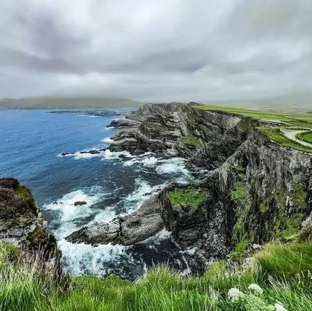 Coastal cliffs with green grassy top, rocky faces, and crashing waves in the Atlantic Ocean under cloudy skies.