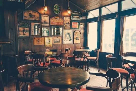 Interior of a cozy bar or restaurant with wooden decor, vintage signs on the wall, and multiple round tables with chairs, near large windows letting in natural light.