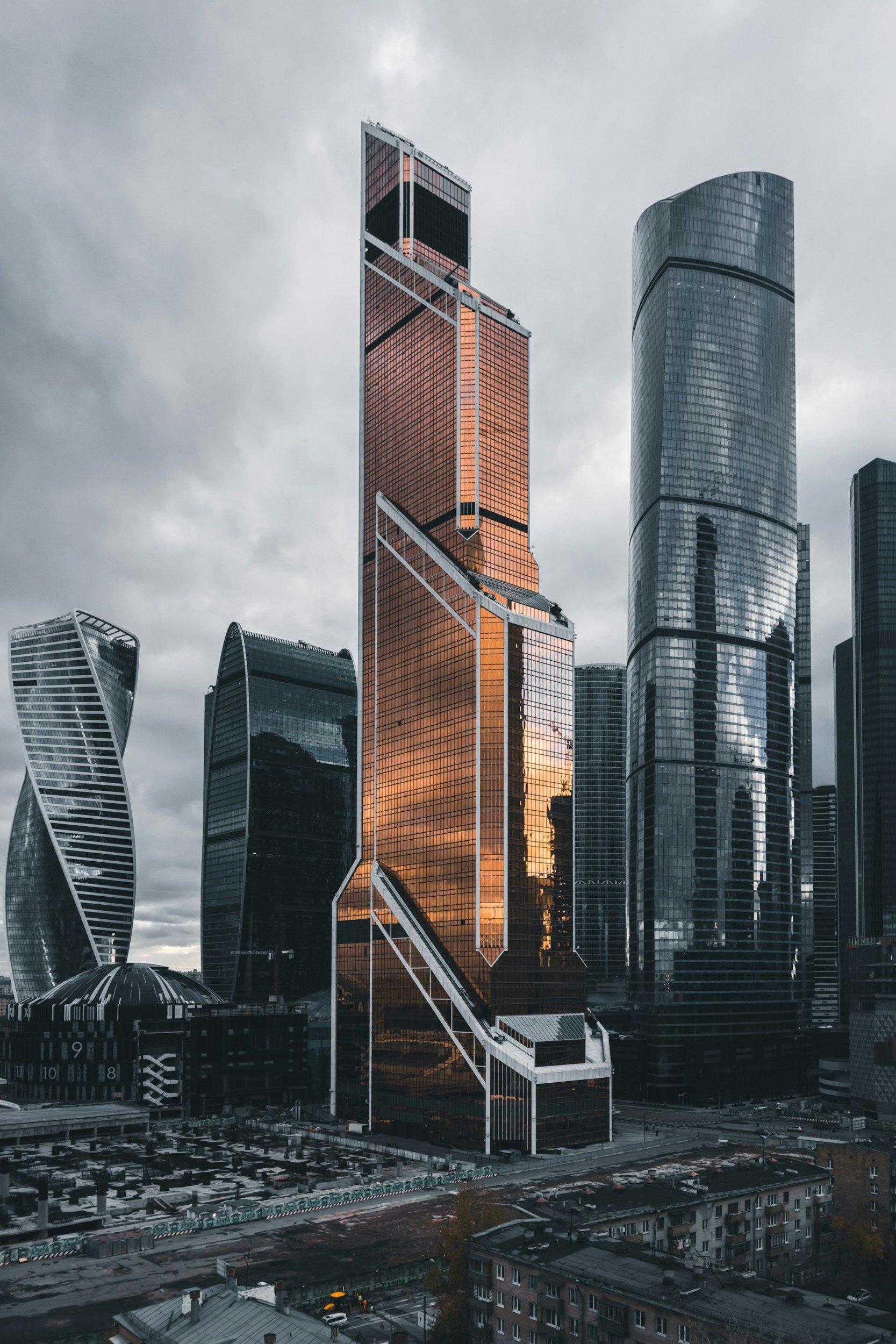 Skyscrapers in a city with dark cloudy skies at sunset, featuring modern glass buildings and a mix of architectural styles.