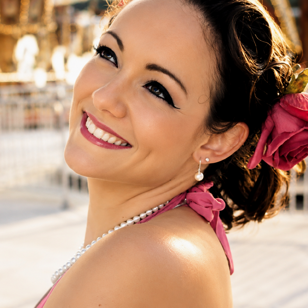 A smiling woman with dark hair styled in curls, wearing pearl earrings, a pearl necklace, and a pink dress with a large pink flower in her hair, outdoors during sunset.