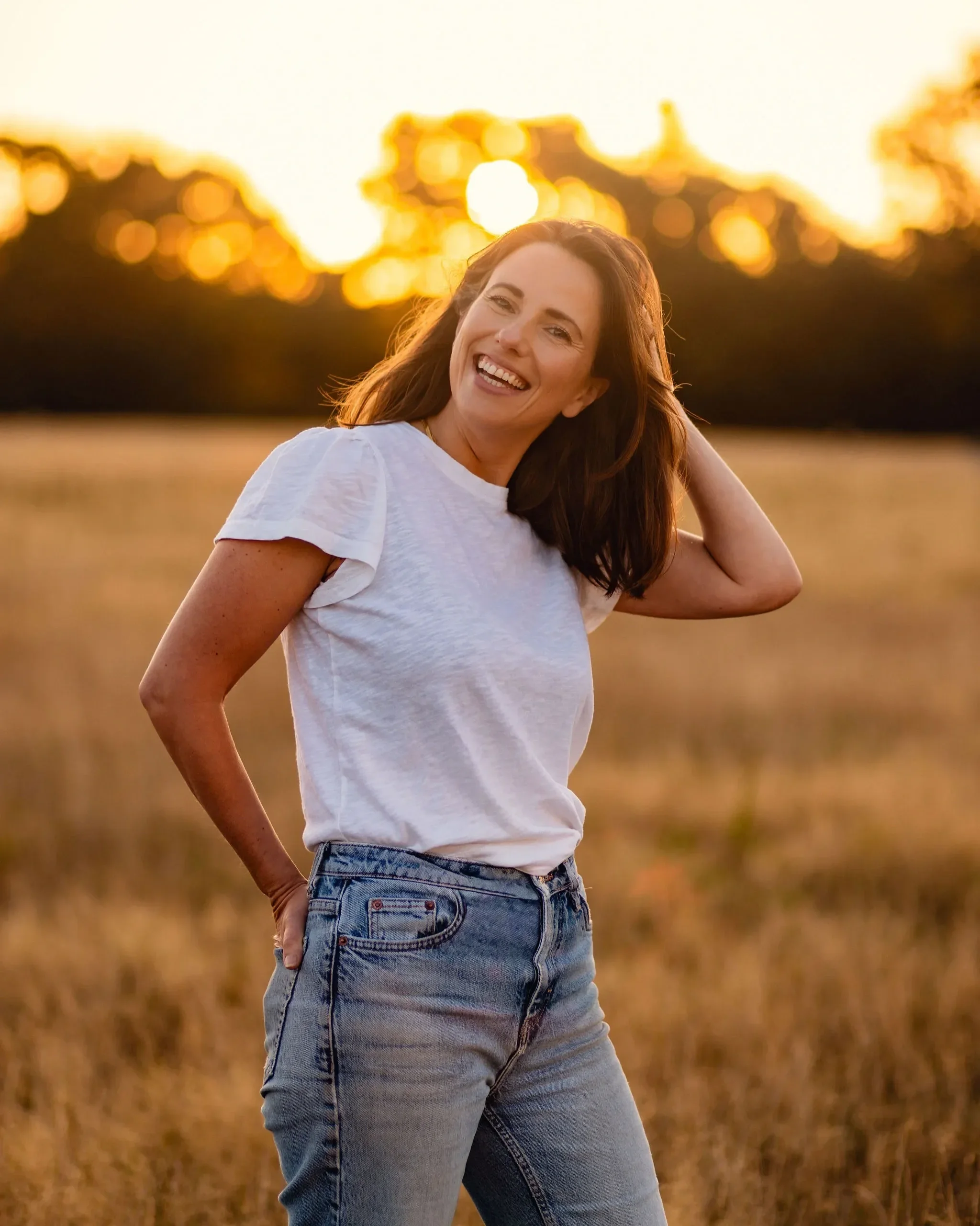 Brunette woman smiling in a sunny field.  She is wearing blue jeans and a white t-shirt