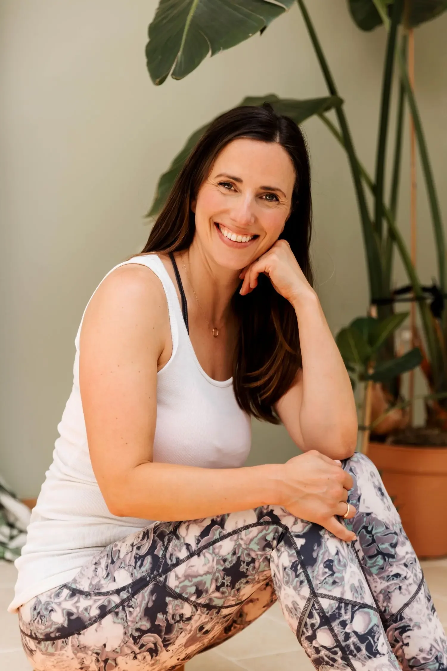 Woman with brunette hair smiling at the camera.  She is sat on the floor, wearing a white vest top and patturned leggings.  There is a tall plant behind her.