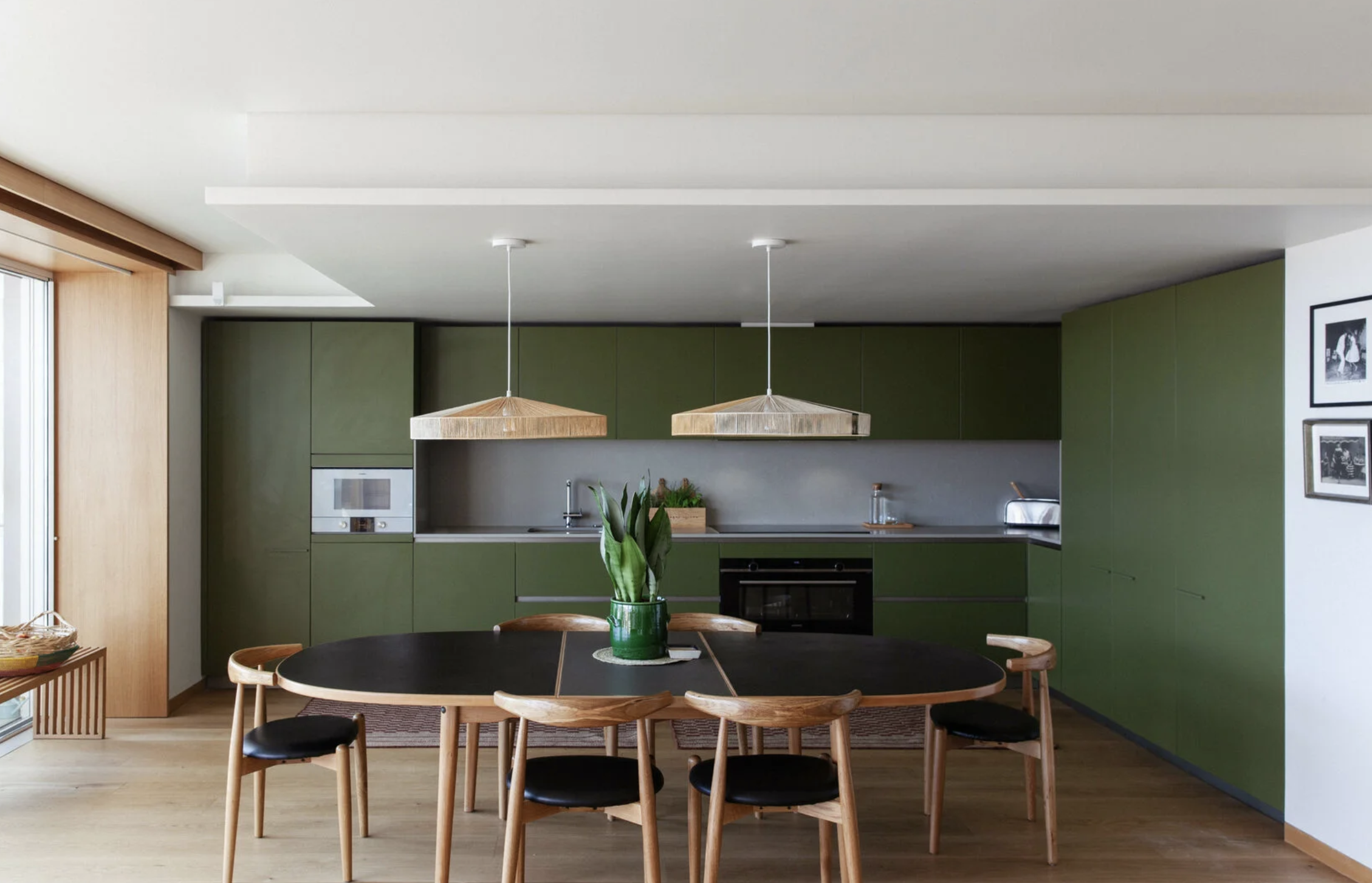 Modern kitchen with green cabinets, wooden round dining table, six black cushioned chairs, hanging wooden pendant lights, potted plant in the center of the table, gray backsplash, and black oven.