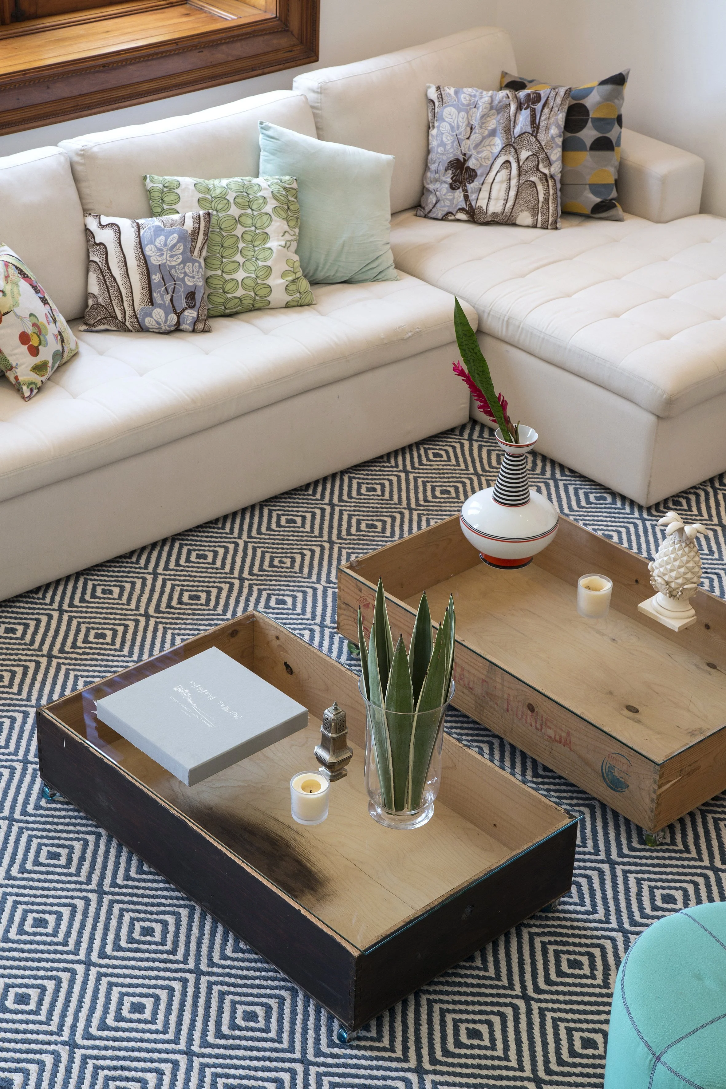 Living room with a white sectional sofa decorated with multiple colorful pillows, a patterned rug, and a wooden coffee table holding a potted plant, candles, a book, and decorative items.