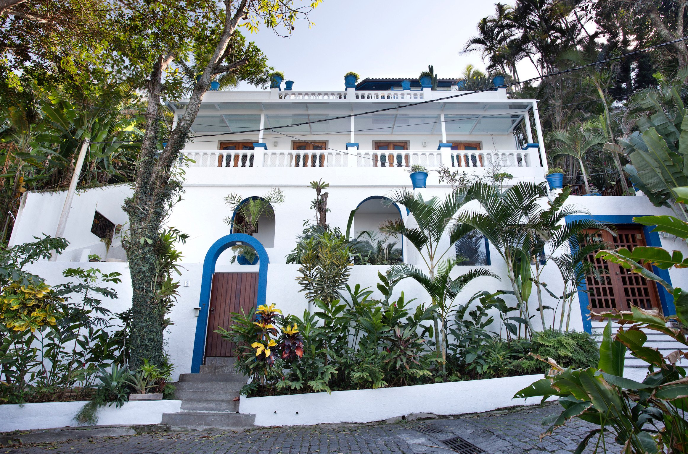 A white multi-story house with blue trim surrounded by lush green tropical plants and trees, with a cobblestone pathway in front.