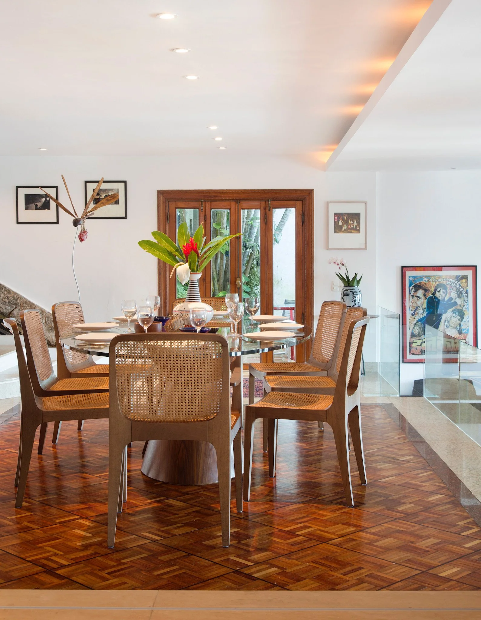 Dining room with a round glass table set for multiple people, wooden chairs with rattan backs, a large floral arrangement on the table, framed pictures on the white walls, a wooden door with glass panes, and hardwood flooring.