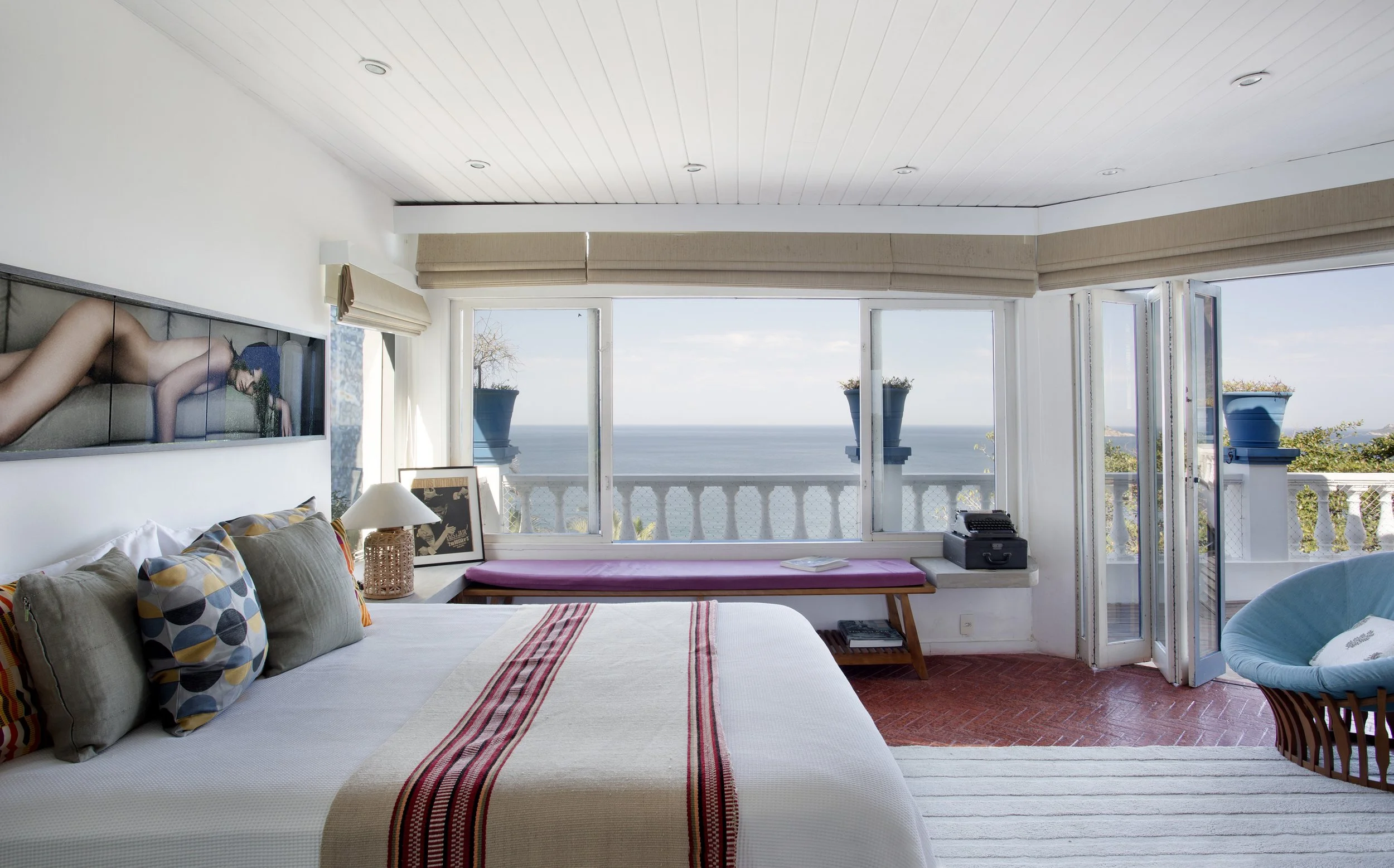 A bedroom with a large bed adorned with multicolored pillows, a framed artwork, a lamp, and a pink window bench. Large windows and a door open to a balcony with a view of the ocean and sky, with potted plants on the balcony.
