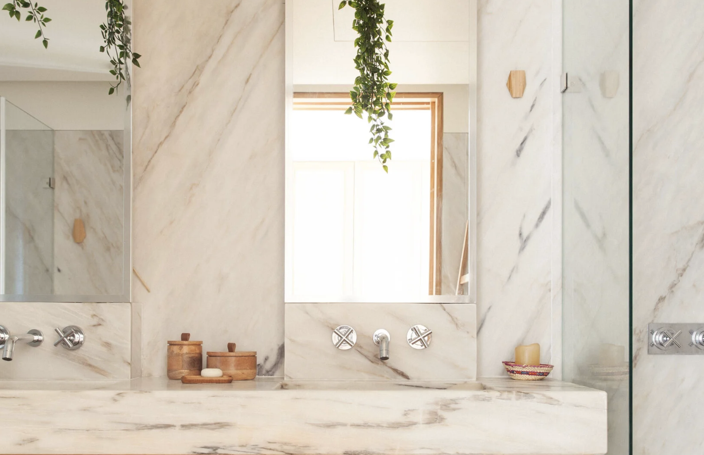 A modern marble bathroom with a large mirror, greenery hanging from the ceiling, and natural light coming through a window. The bathroom counter has wooden containers and a soap dispenser, with a colorful basket on the side.