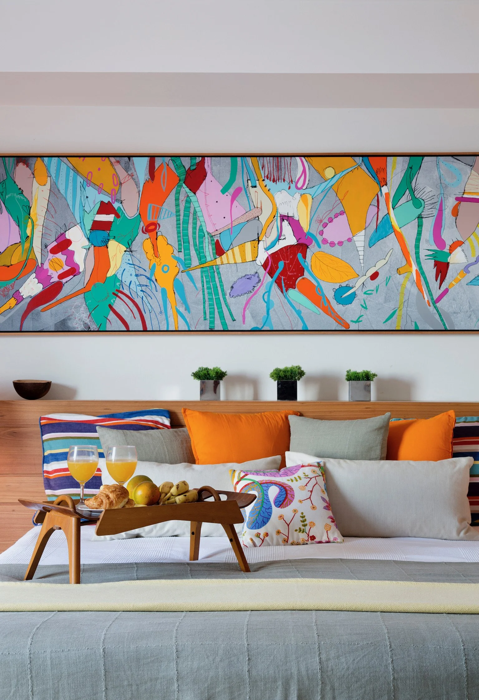 Living room with a beige sofa decorated with colorful pillows and a light gray blanket, a wooden tray with breakfast on a bed, an abstract colorful painting on the wall, three small potted plants, and two glasses of orange juice on the tray.