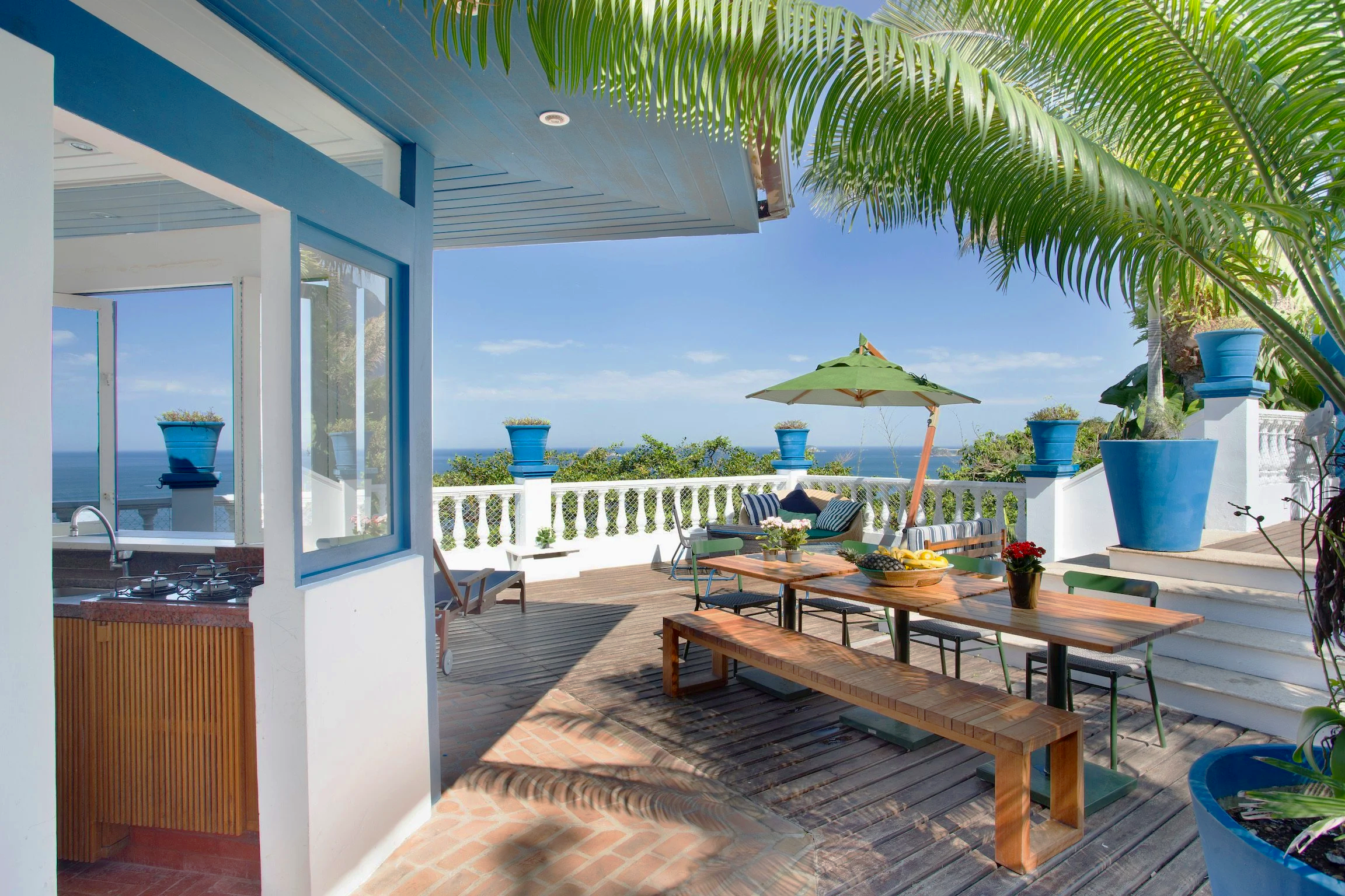 An outdoor patio area with a wooden dining table, chairs, large blue flower pots, and a green umbrella. There are views of the ocean and sky in the background, with tropical plants including palm trees.