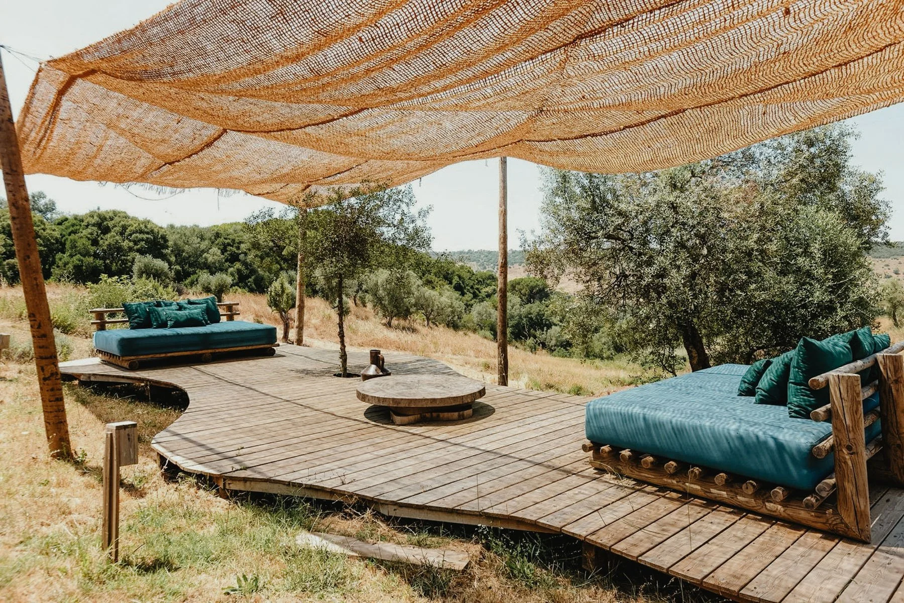 Outdoor seating area with a wooden deck, two blue cushioned lounge chairs or daybeds with green pillows, a round stone table, and a rustic shade canopy made of woven fabric, set in a scenic landscape with trees and hills in the background.