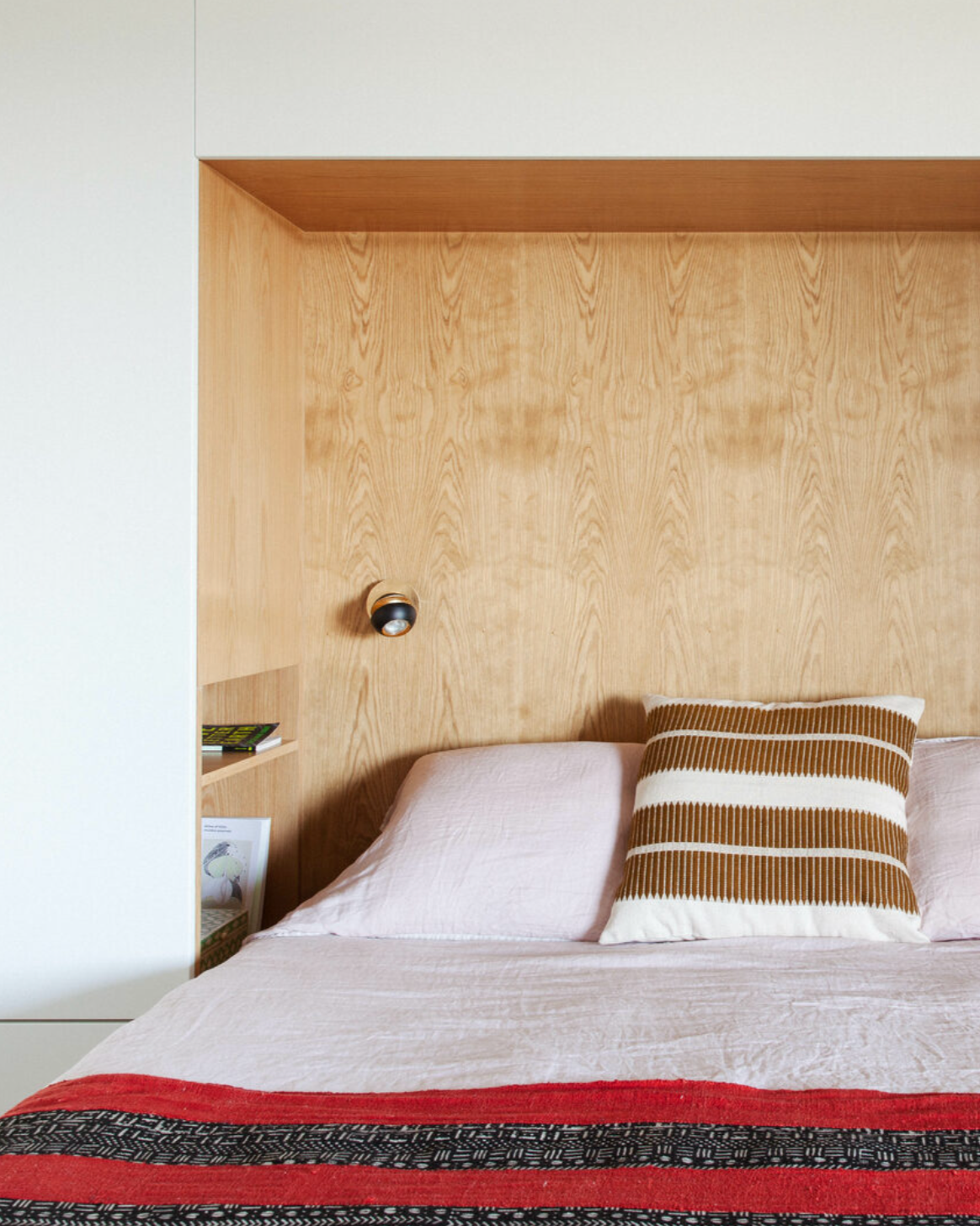 A bedroom with a wooden headboard, a bed with pink sheets, a striped pillow, a red and black patterned blanket, a small side shelf with books and framed art, and a wall-mounted reading light.
