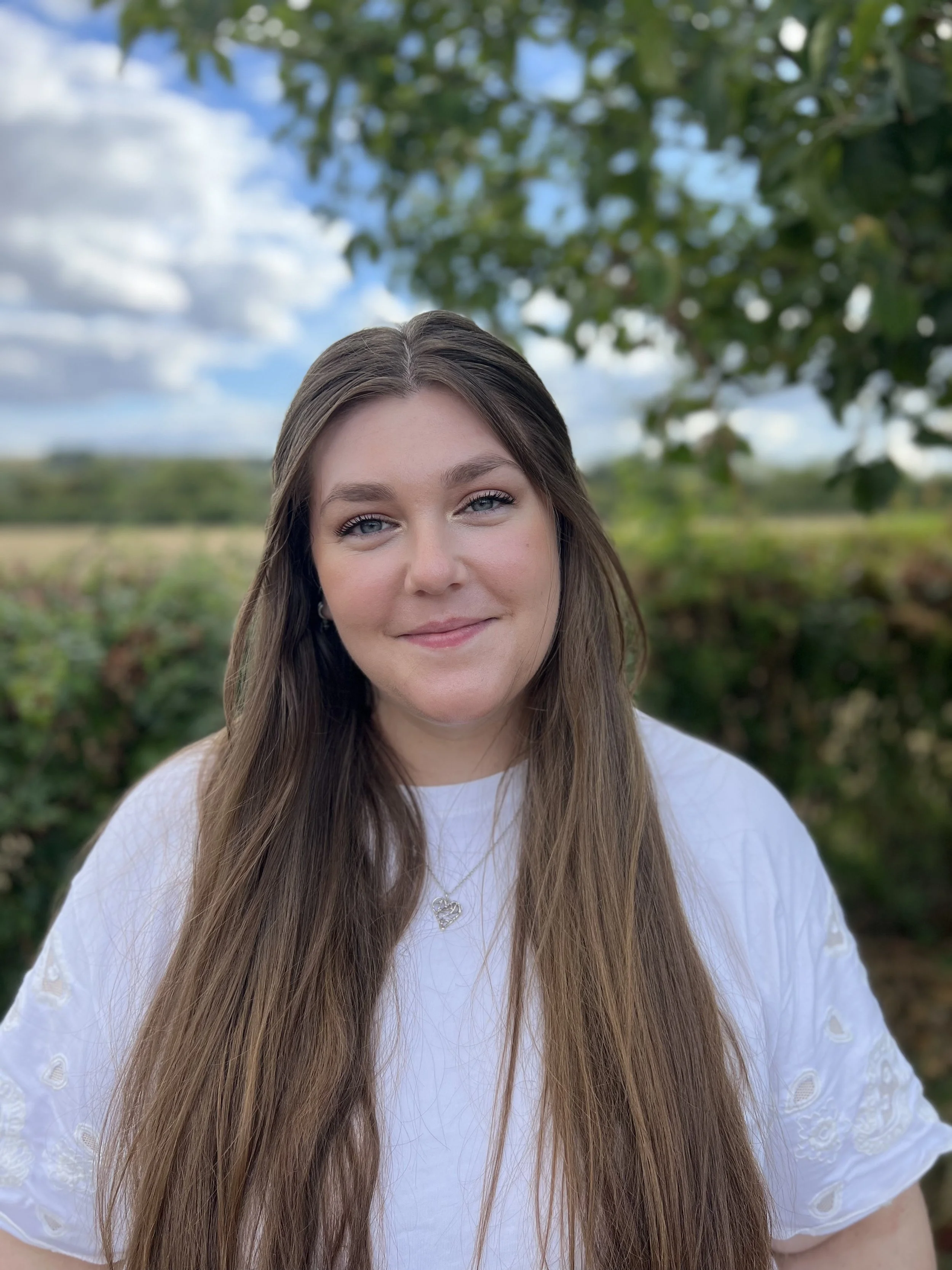 A female therapist with long, light brown hair standing outdoors with a blue sky, white clouds, and green trees in the background.