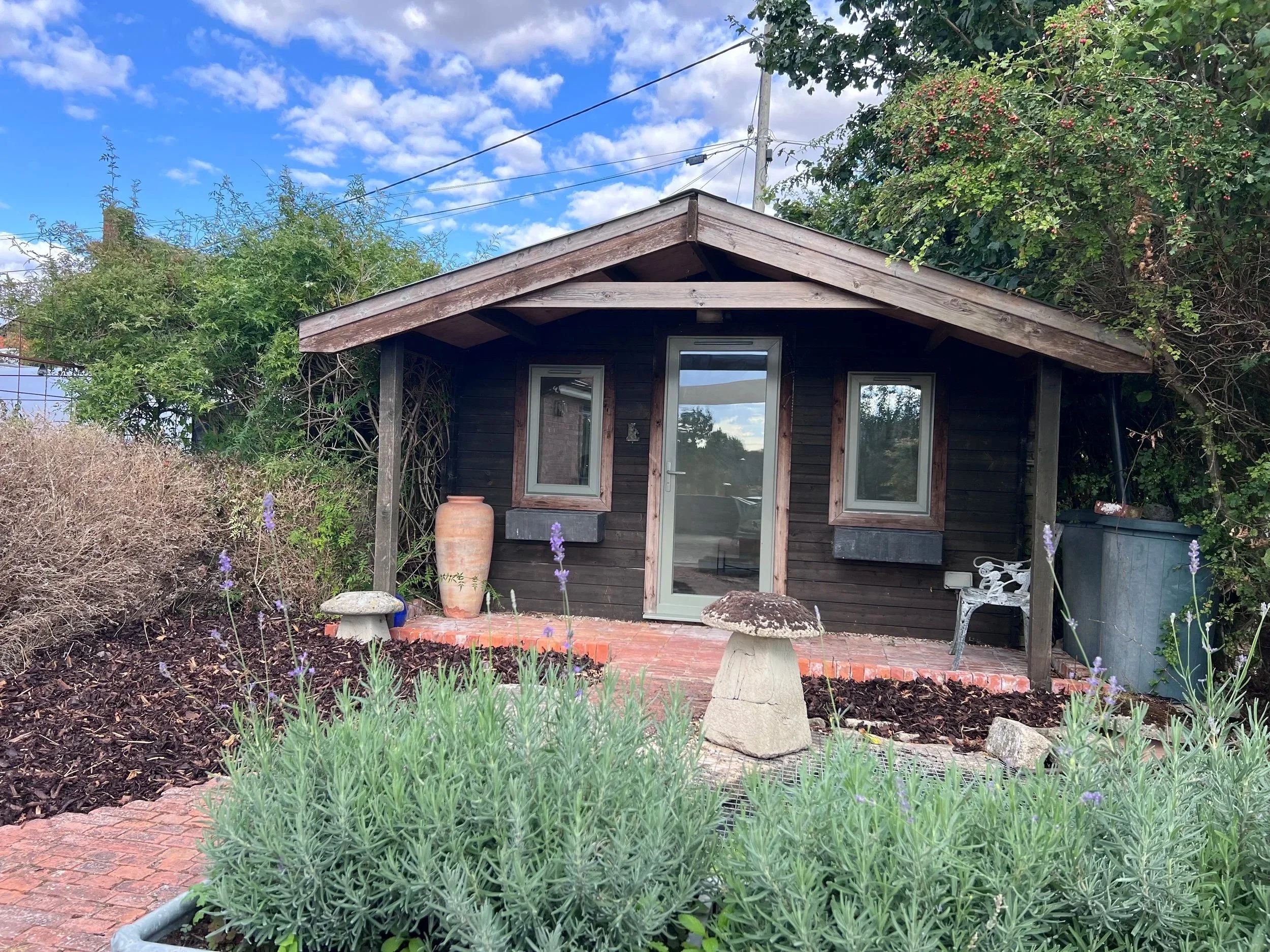 A small wooden therapy room near Devizes and Calne with a glass door and two windows, surrounded by a garden with green plants, purple flowers, and decorative stones, with a clear blue sky and fluffy white clouds above.