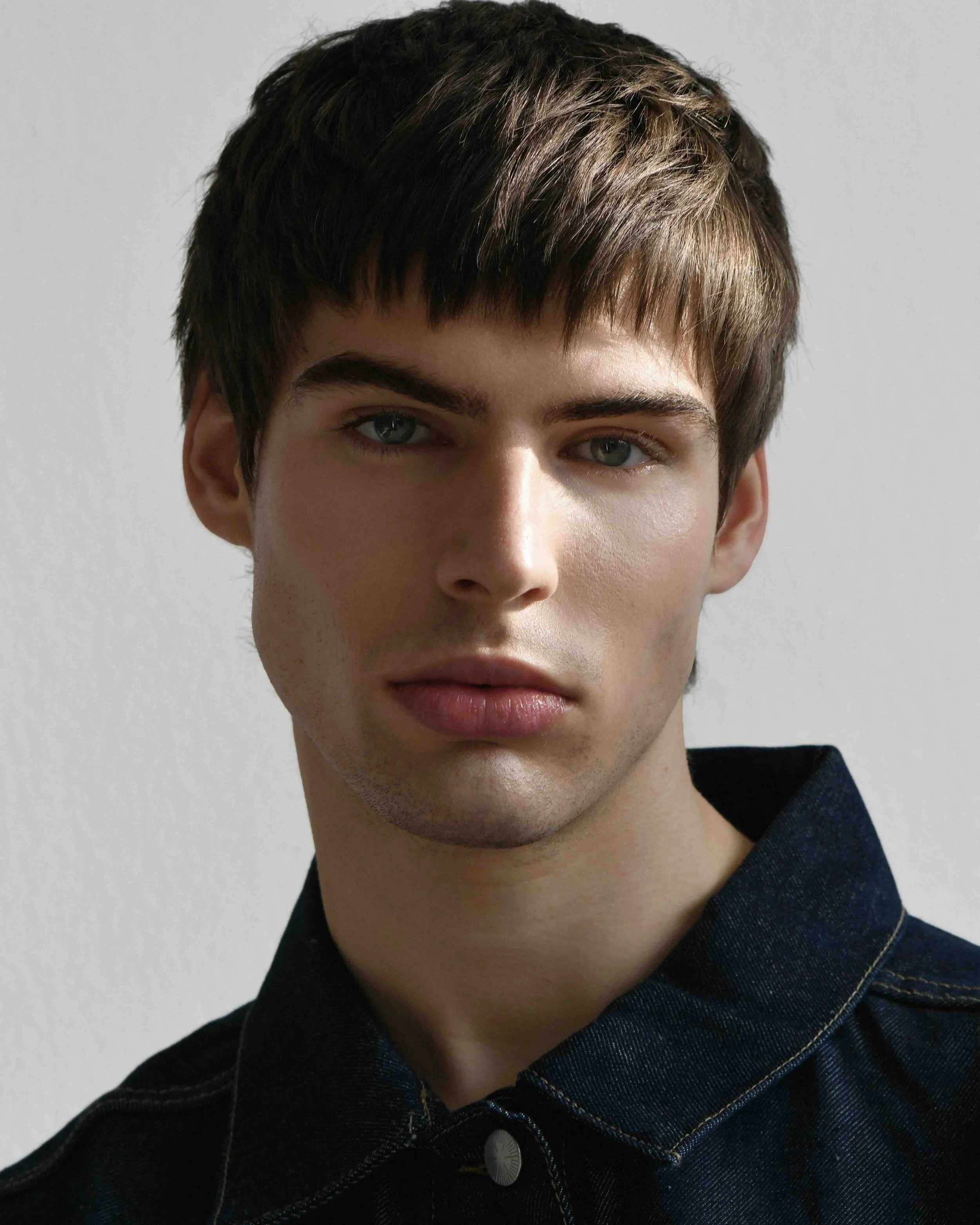 Nahaufnahme eines jungen Mannes mit kurzen braunen Haaren, blauen Augen, hellhäutiger Haut und schwarzen Hemd mit sichtbar gesticktem Muster.