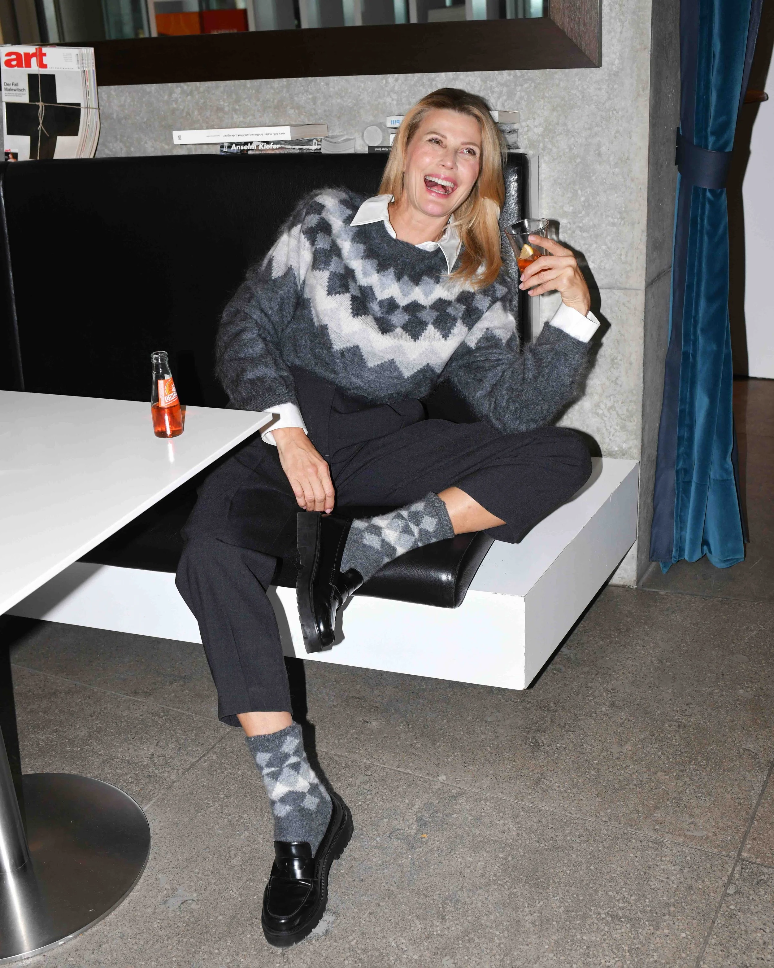 Eine Frau sitzt auf einer schwarzen Lehne und ist am Lachen. Sie trägt einen grauen Pullover mit einem weißen Hemd darunter, schwarze Hosen, schwarze Schuhe, graue Socken mit Mustern und hält ein Glas mit einem Getränk. Vor ihr auf einem weißen Tisch steht eine kleine rote Flasche. Im Hintergrund sind Bücher und Zeitschriften, und es gibt einen blauen Vorhang auf der rechten Seite.