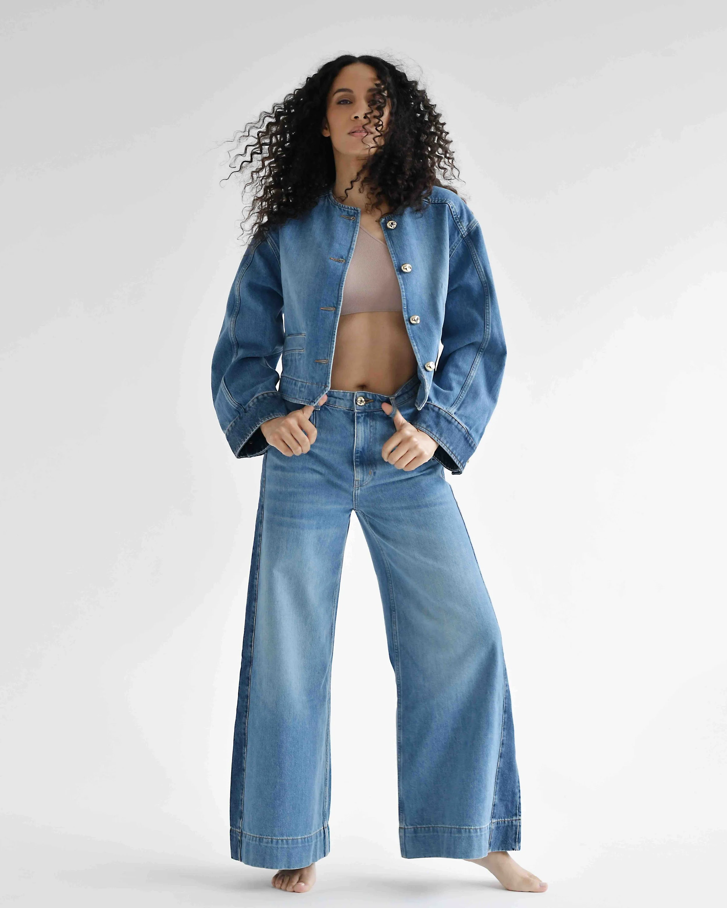 Eine Frau trägt ein Denim-Jacket und -Hose, die Hose ist sehr breit und lang, sodass ihre Füße nur teilweise sichtbar sind.