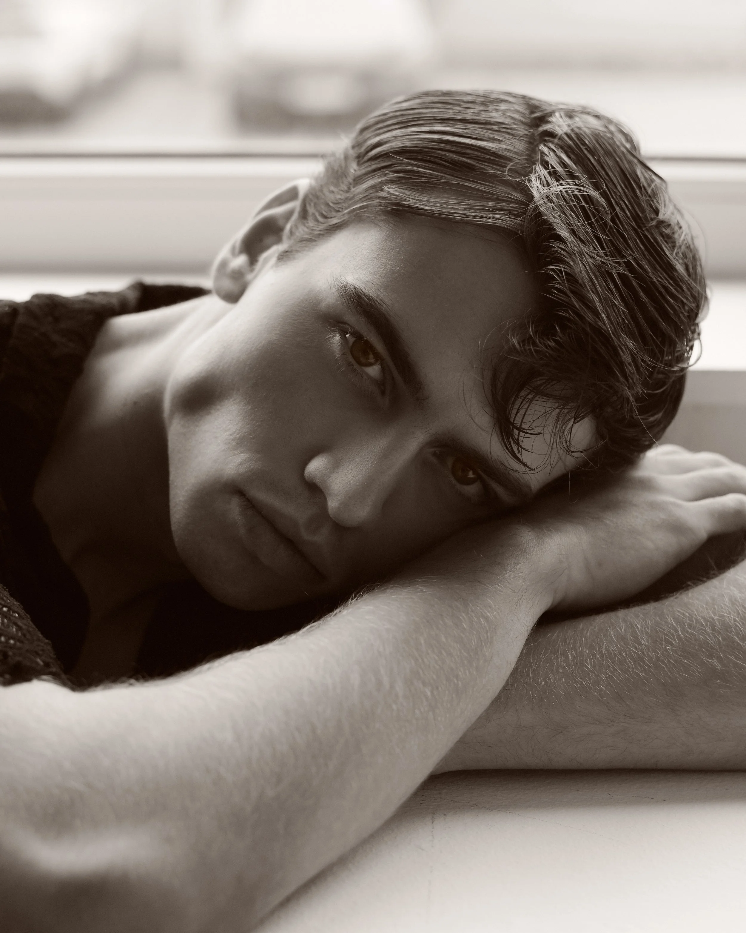 Schwarz-Weiß-Fotografie eines jungen Mannes, der flach auf einem weißen Kissen liegt, mit seinem Kopf seitlich nach oben geneigt, Blick in die Kamera, kurze, gepflegte Haare, entspannte Ausstrahlung.