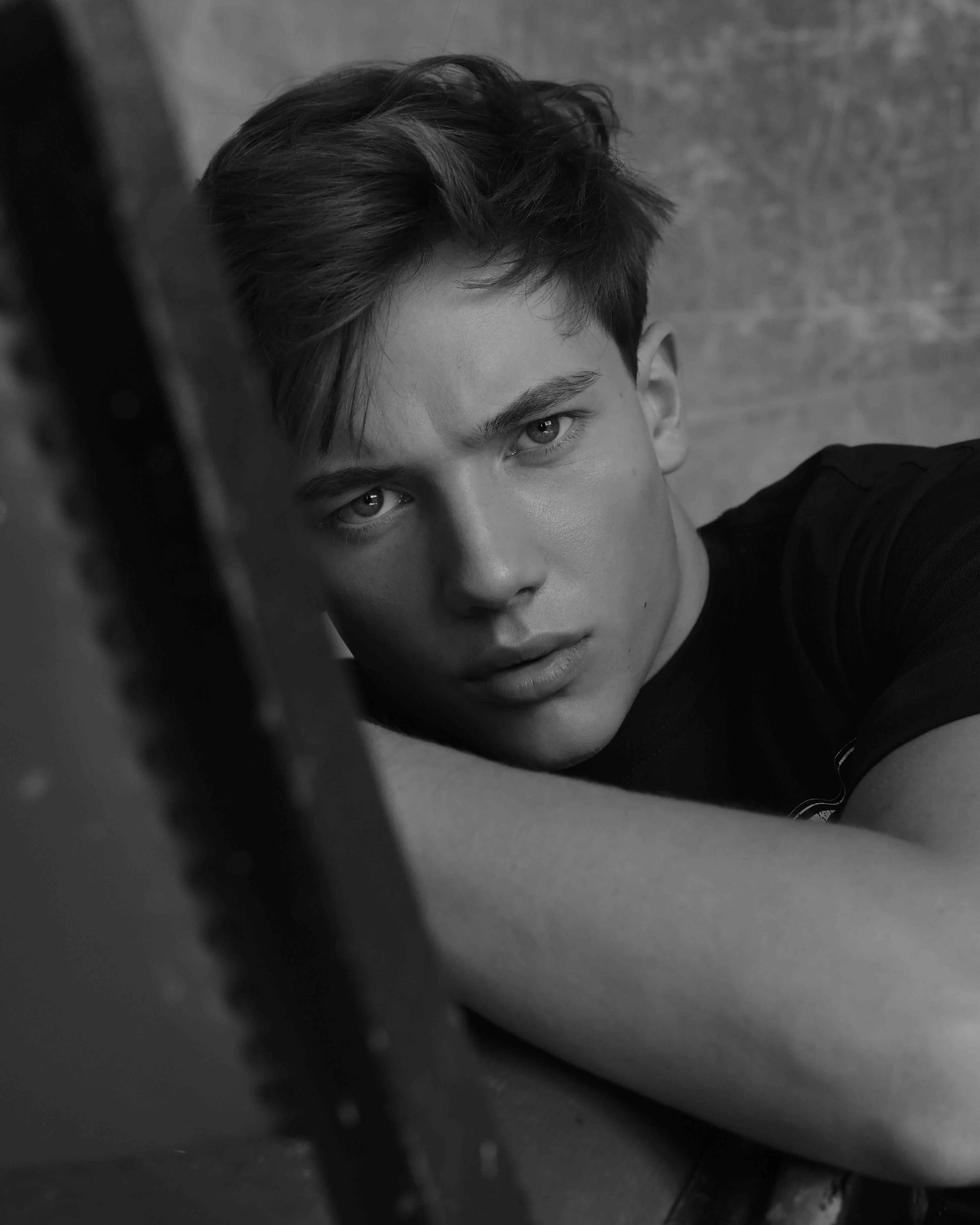Schwarz-weiß-Foto eines jungen Mannes mit kurzen, gestylten Haaren, der direkt in die Kamera schaut. Er befindet sich in einer Nahaufnahme, angeschnitten an der Schulter, mit einem grauen Hintergrund.