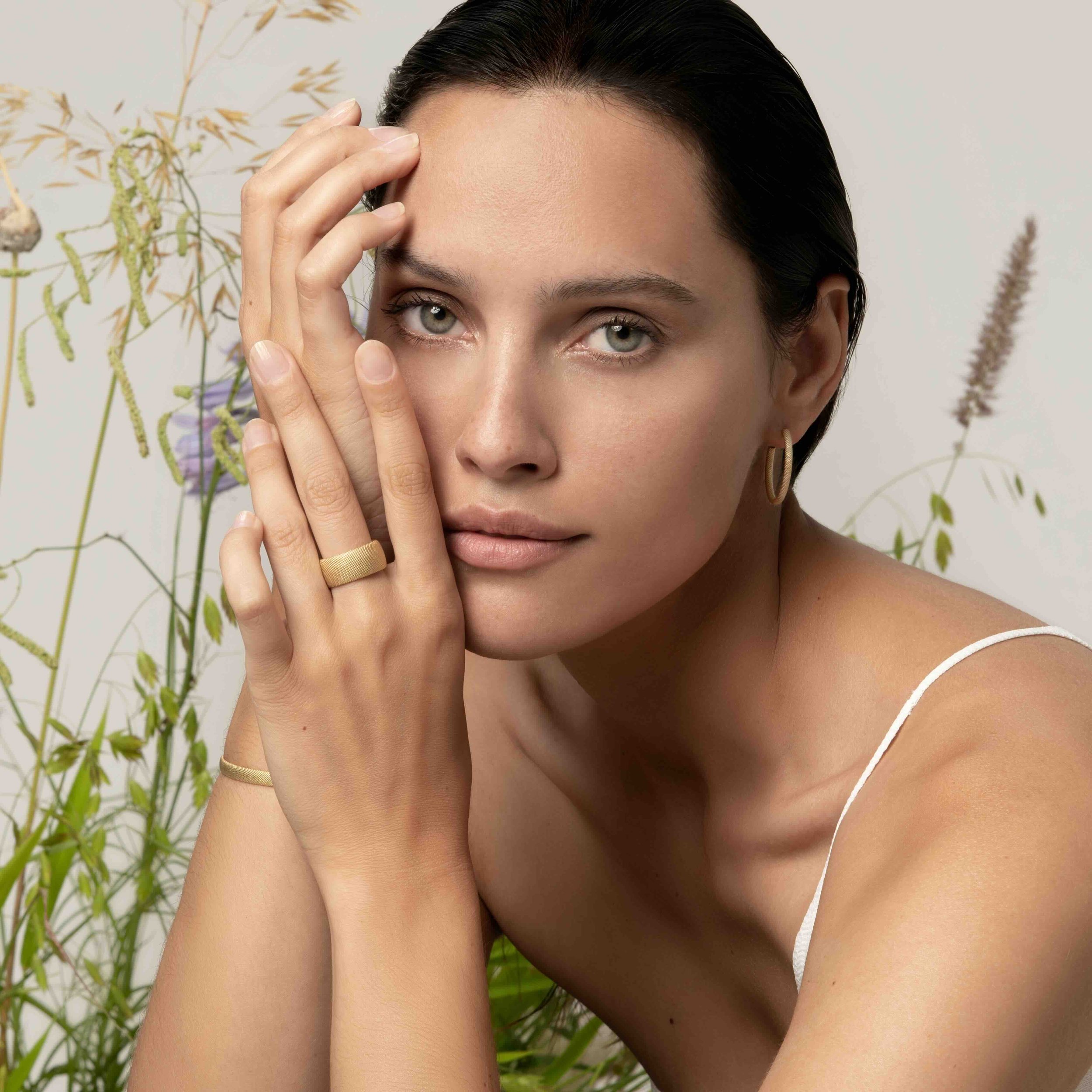 Nahaufnahme eines jungen Frauen, die ihr Gesicht halb verdeckt mit der Hand hält, mit dunkelbraunen Haaren und blauen Augen, vor einem neutralen Hintergrund mit einigen grünen und lila Pflanzen.