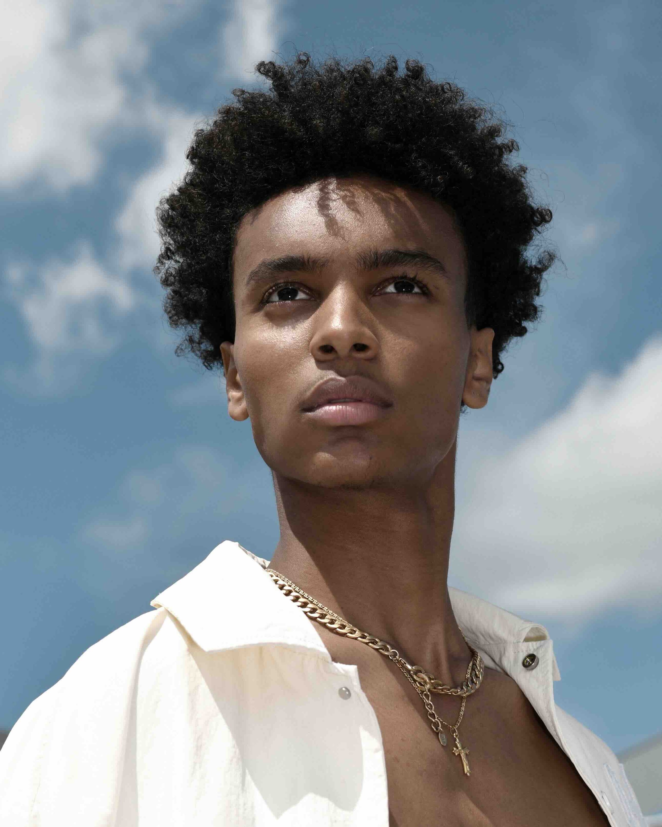 Nahaufnahme eines jungen Mannes mit lockigem schwarzen Haar vor einem blauen Himmel mit Wolken, trägt eine weiße Jacke, goldene Halsketten.