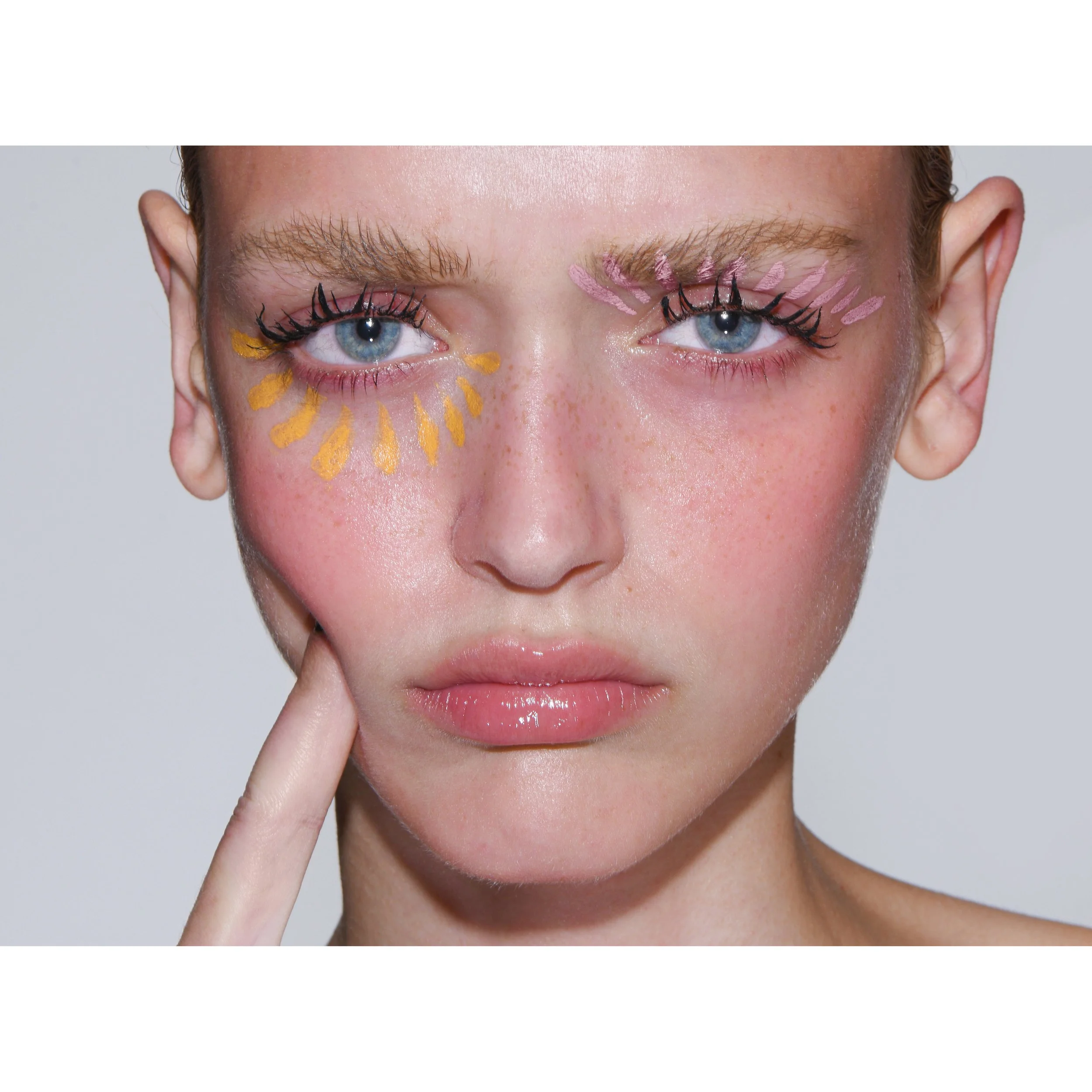 Nahaufnahme eines Gesichts mit blondem Haar und blauen Augen, mit buntem Make-up um die Augen, inklusive schwarzer Wimperntusche und Pastellfarben auf den Lidern, das aufhellende Hautbild und eine Person, die in die Kamera schaut.