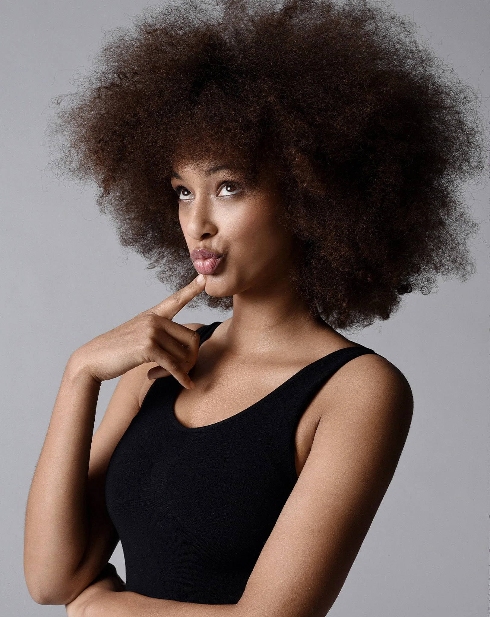 Junge Frau mit lockigem Afro-Haar, die den Finger an die Lippen legt und nachdenklich schaut, vor grauem Hintergrund, trägt schwarzes Oberteil.