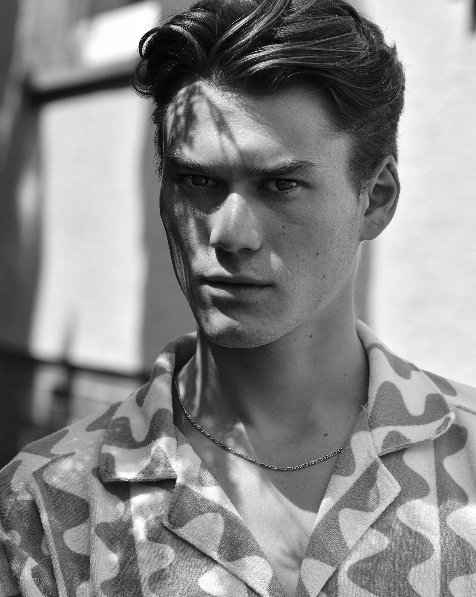 Porträt eines jungen Mannes mit dunklen Haaren, sichtbarem Hautbild, in Schwarz-Weiß, trägt eine Joggingjacke mit Camouflage-Muster und eine Silberkette.
