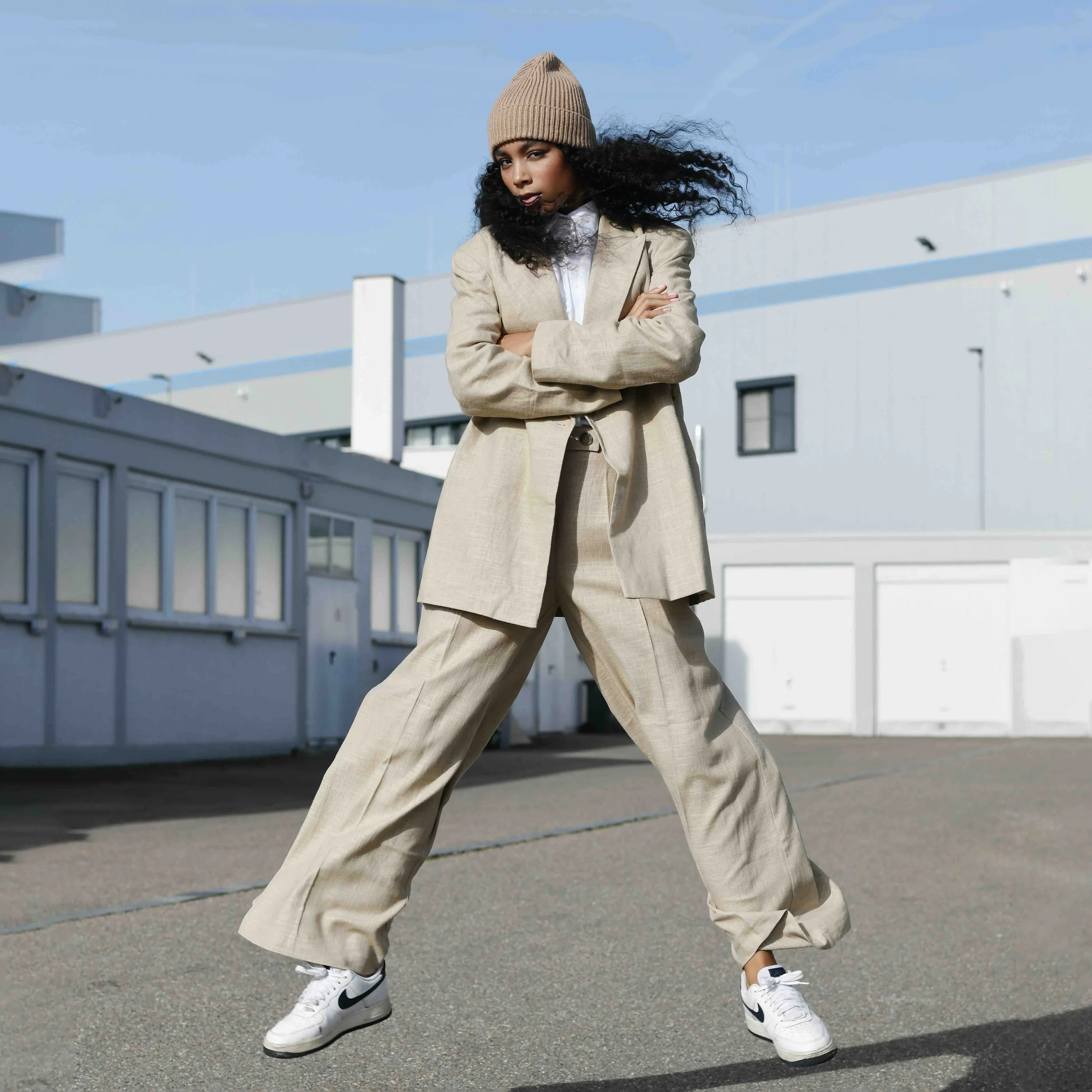 Junge Frau trägt beige Anzug, beige Mütze, weiße Nike Sneaker, steht selbstbewusst mit verschränkten Armen vor einer modernen weißen Gebäudefassade unter blauem Himmel.