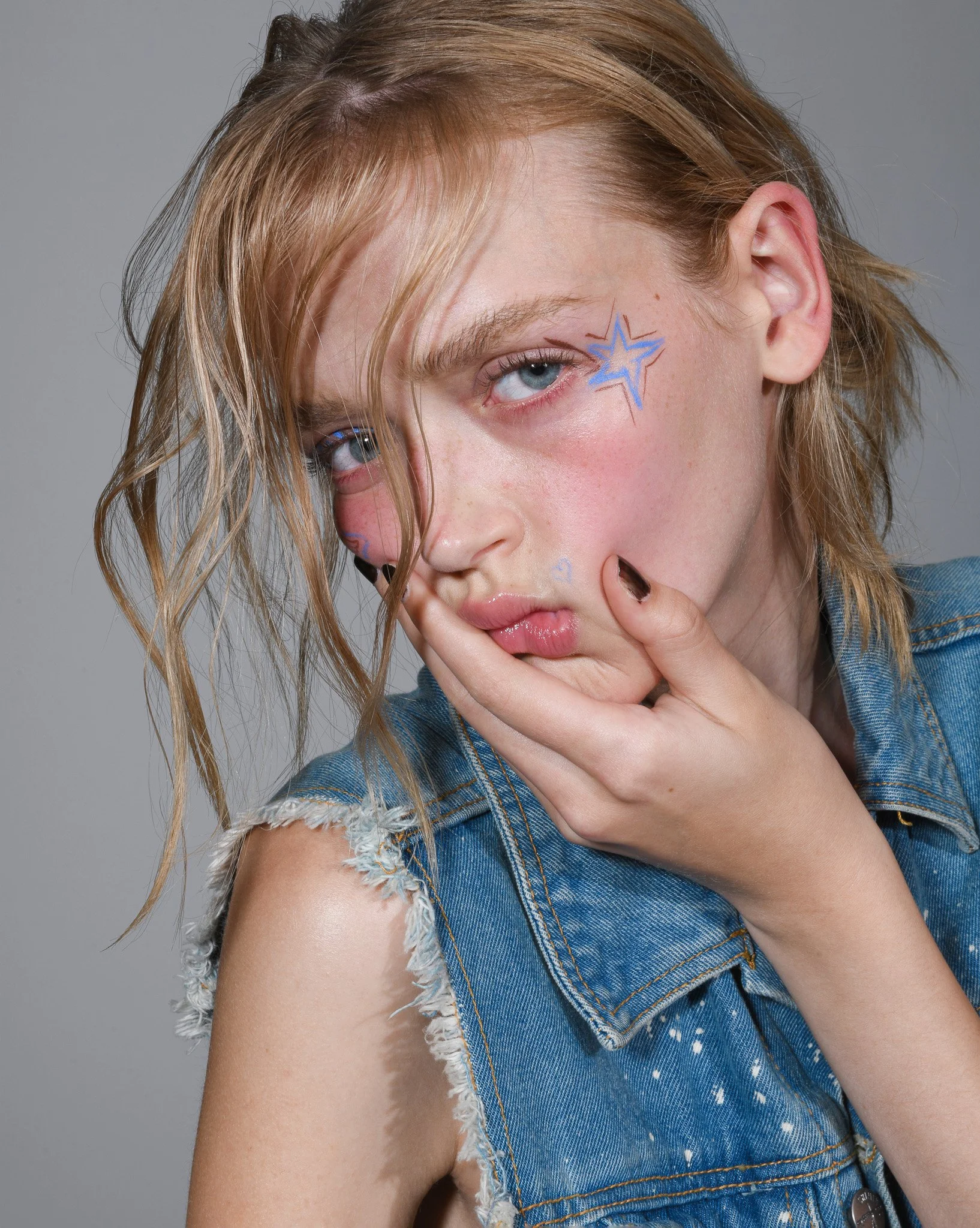 Nahaufnahme eines jungen Mädchens mit blonden, lockigen Haaren, die mit blauer Gesichtszeichnung in Form eines Sterns verziert sind, trägt eine abgenutzte Blue-Jeansjacke mit offenen Schultern, die auf grauem Hintergrund liegt.