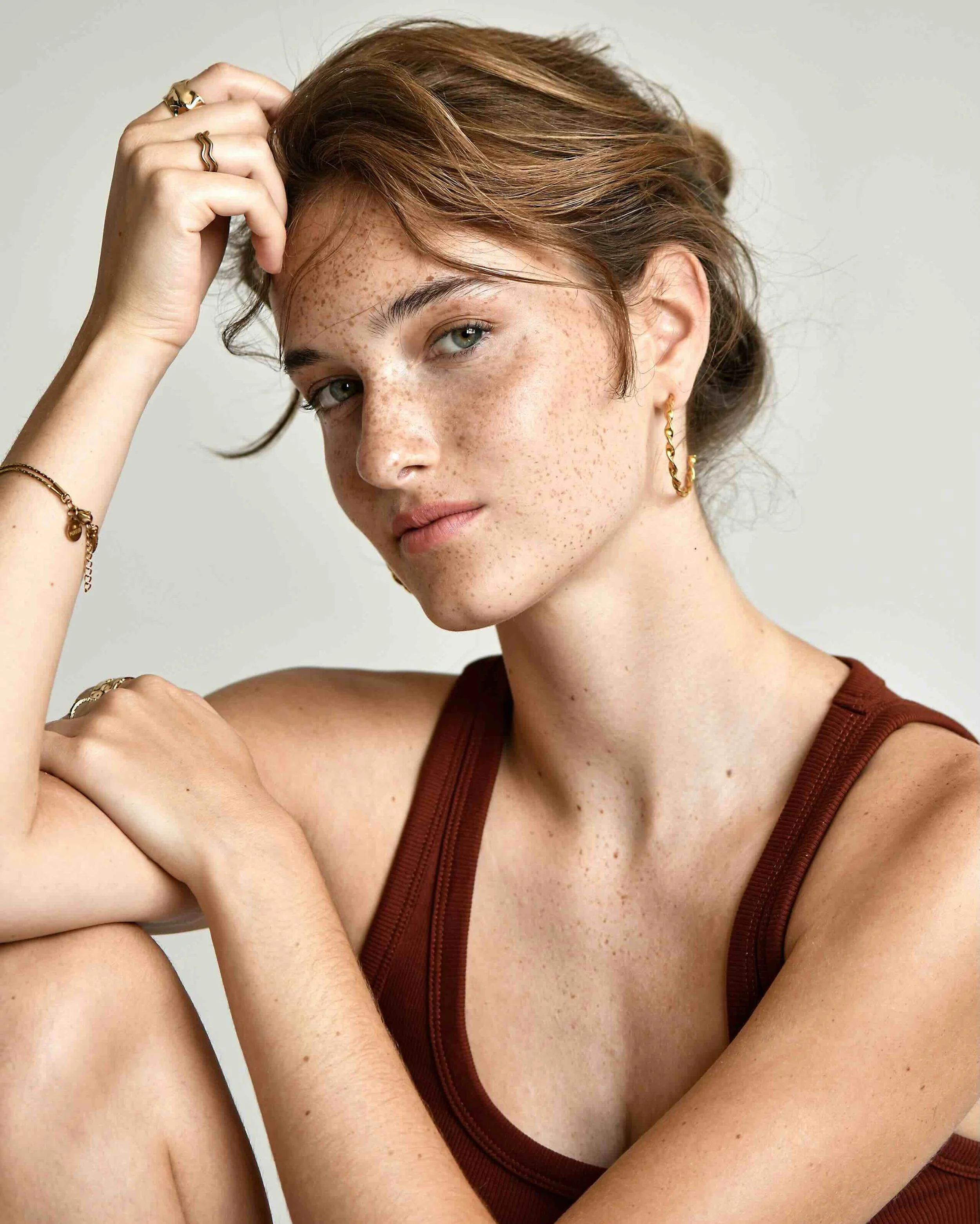Ein junges Mädchen mit sommersprossiger Haut, braunen kurzen Haaren und Goldschmuck am Ohr und Hand, trägt ein braunes Tanktop, sitzt lässig und schaut nachdenklich.