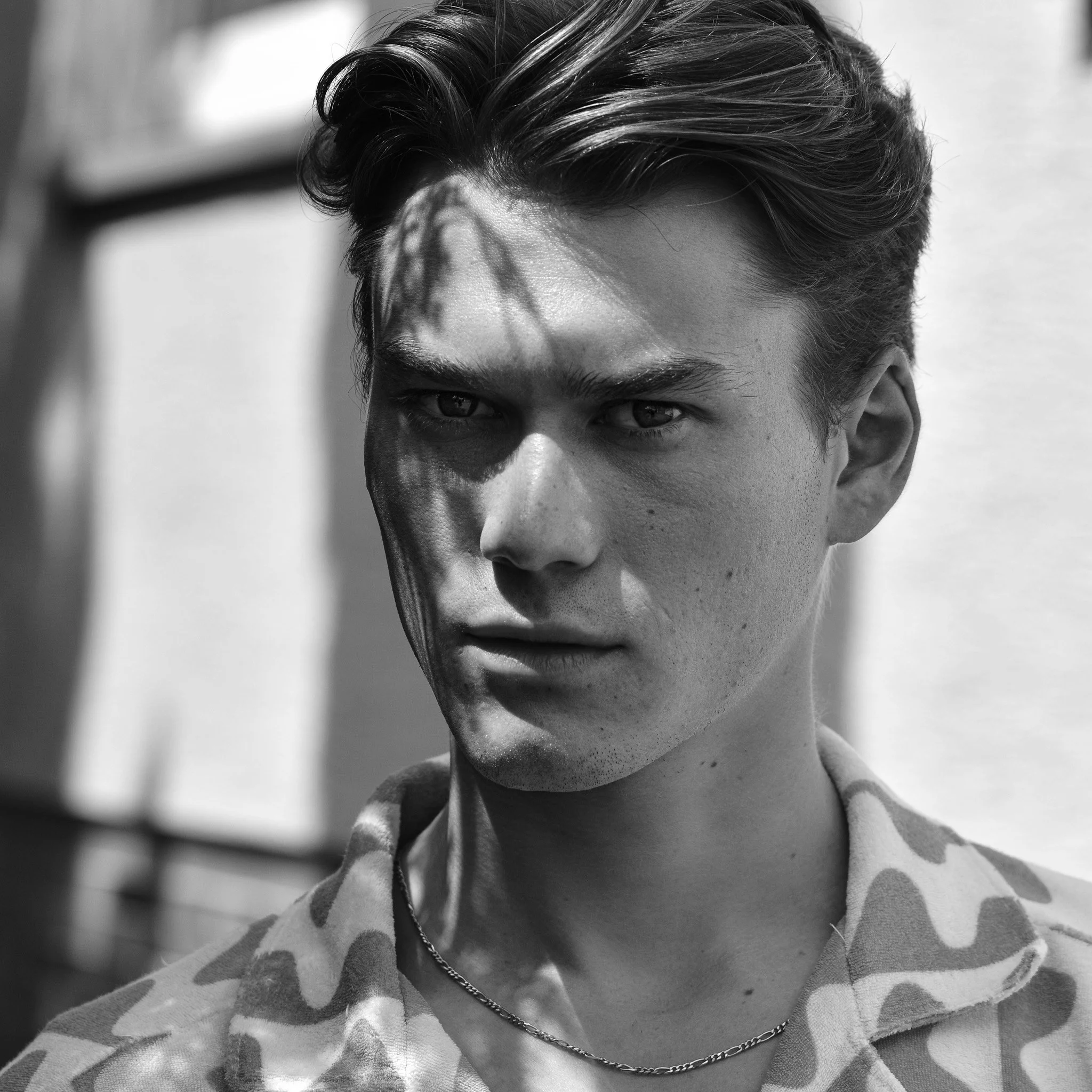 Schwarz-weiß-Foto eines jungen Mannes mit dunklem, gewelltem Haar, sichtbar nur der Teil seines Gesichts und der Schulter, im Hintergrund unscharf, mit Sonnenlicht und Schatten.