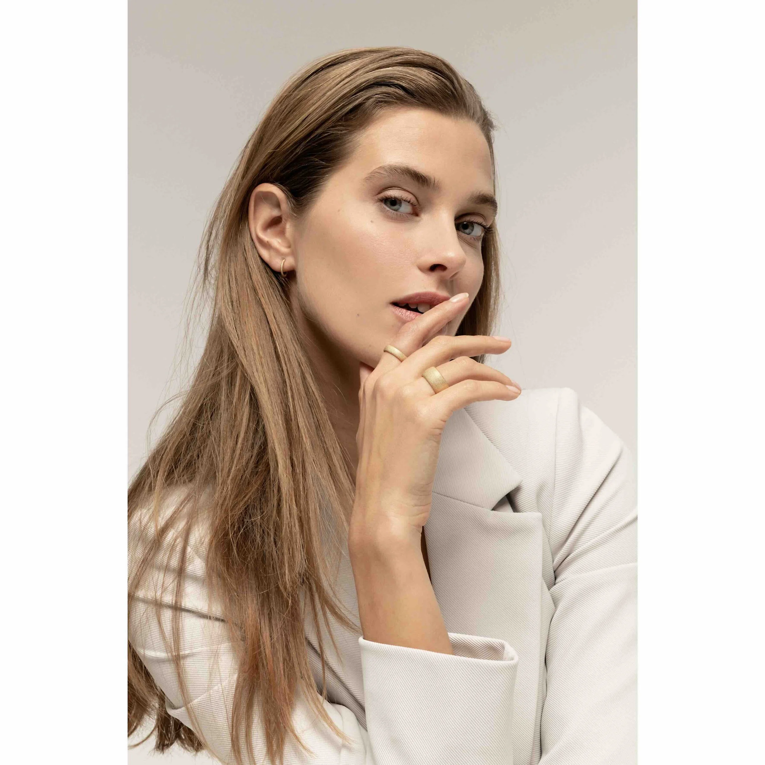 Nahaufnahme einer jungen Frau mit langem, braunem Haar, hellen Augen, dezentem Make-up, goldenen Ohrringen, einem Ring am Finger und einem weißen Blazer vor hellem Hintergrund.