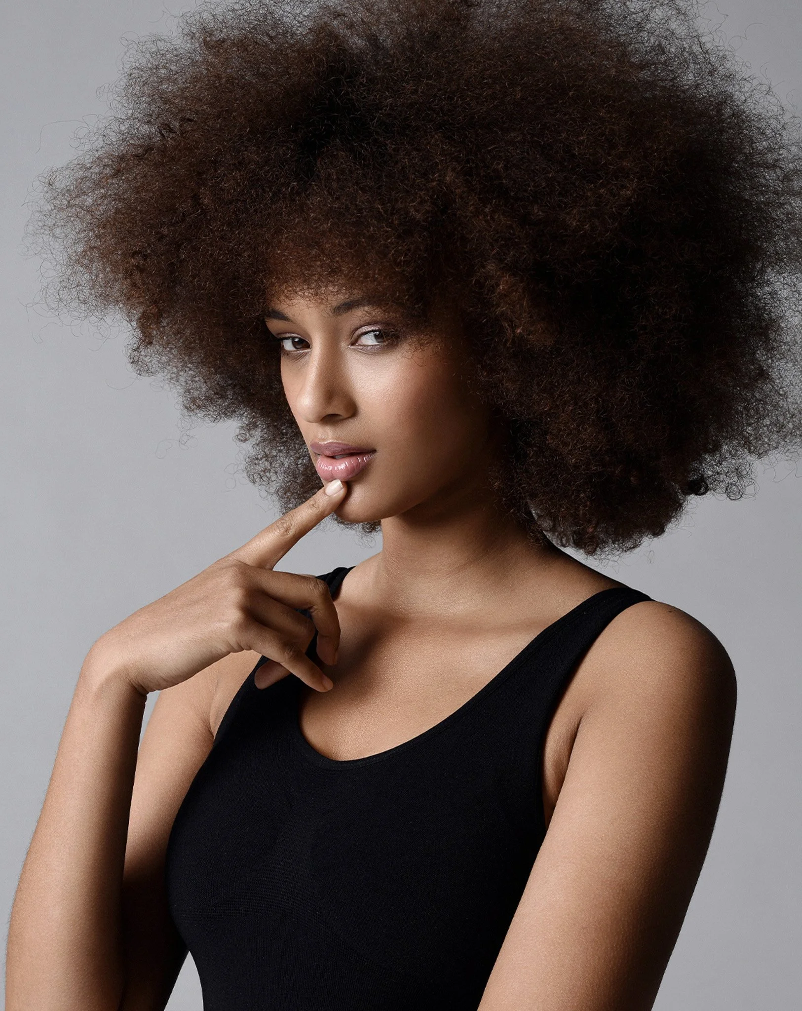 Porträt einer dunkelhäutigen Frau mit lockigem Afro-Haar, die vor einem grauen Hintergrund steht, in schwarzem Tanktop, nachdenklich, mit dem Finger am Kinn.