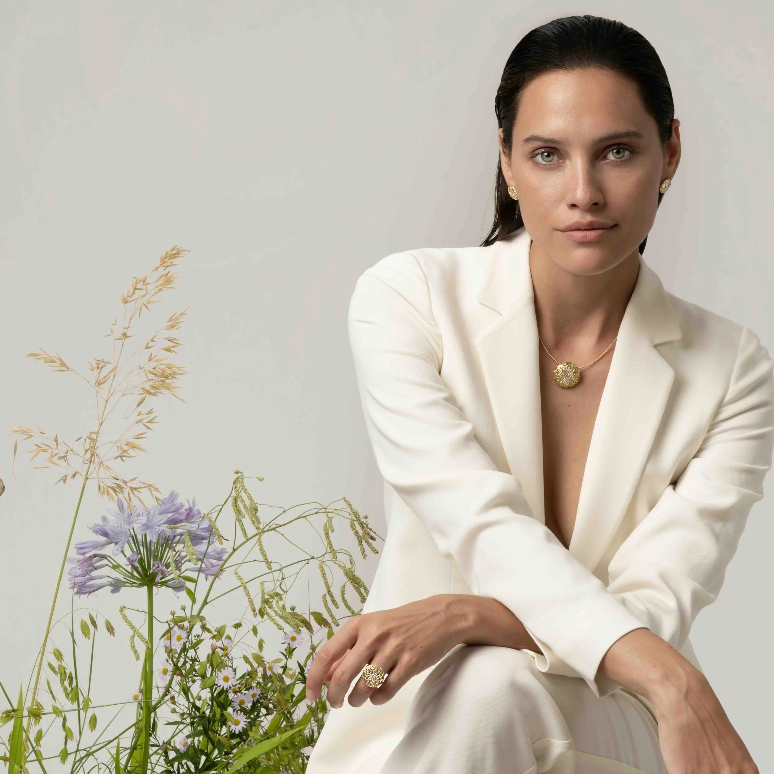 Eine Frau in einem weißen Blazer trägt goldene Ohrringe und einen passenden Halsreif. Sie sitzt vor einer neutralen weißen Wand, während im Vordergrund Blumen und Gräser sichtbar sind.