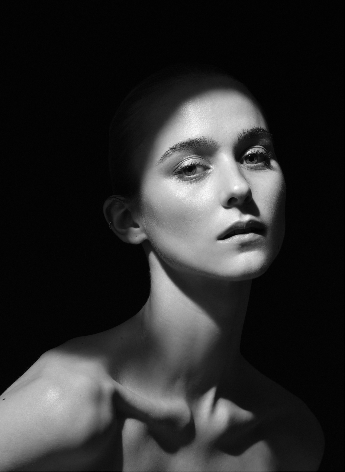 Schwarz-weiß-Porträt einer jungen Frau mit glatter, lichtreflektierender Haut, neugierigen Blick und kurzen, hochgesteckten Haaren, vor dunklem Hintergrund.
