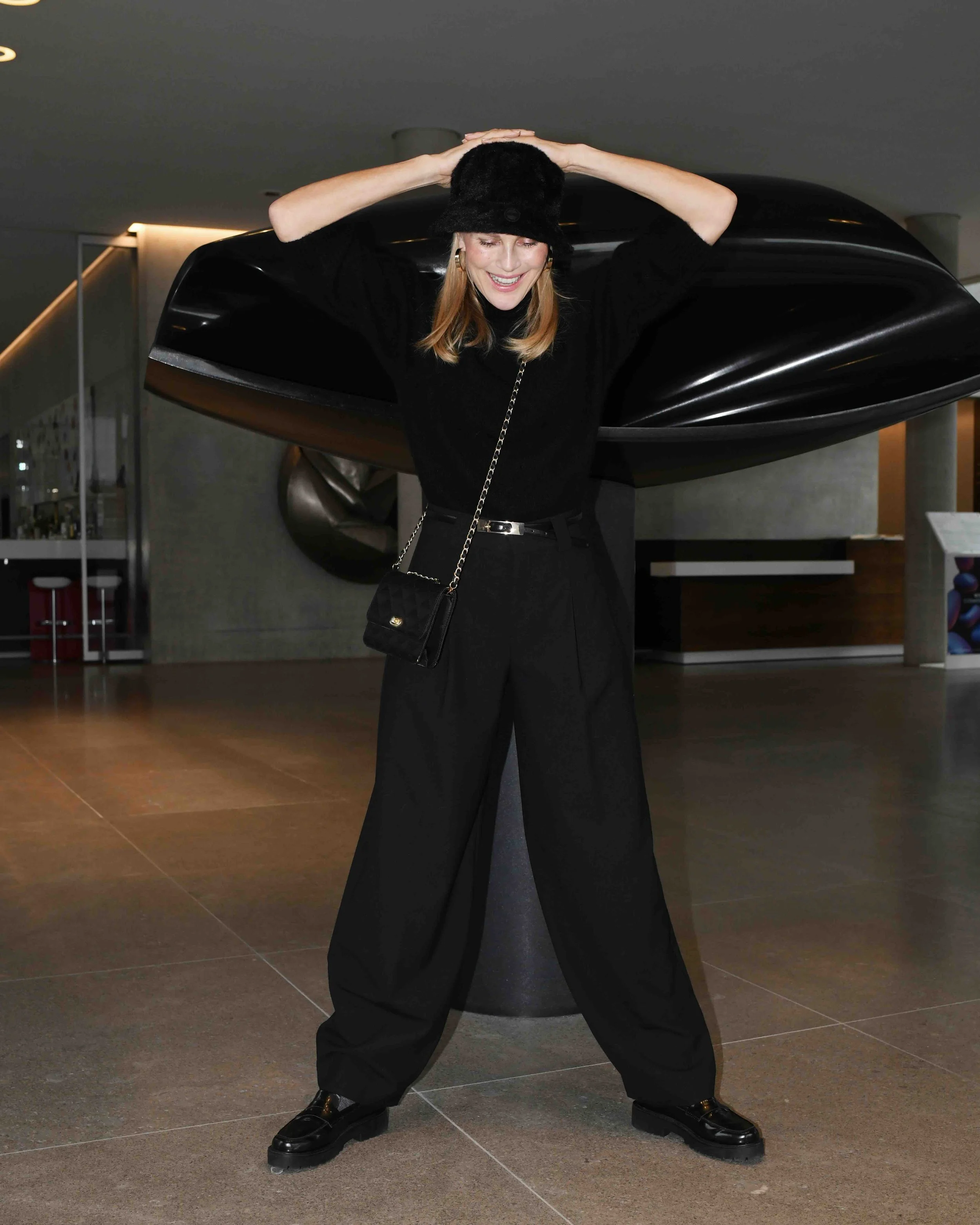 Eine lächelnde Frau mit einem schwarzen Hut und wegen ihres schwarzen Outfits, Schwarz kombiniert mit Gold Accessoires, steht in einer modernen, gut beleuchteten Innenraum, vermutlich einer Kunsthalle, vor einer abstrakten schwarzen Skulptur.