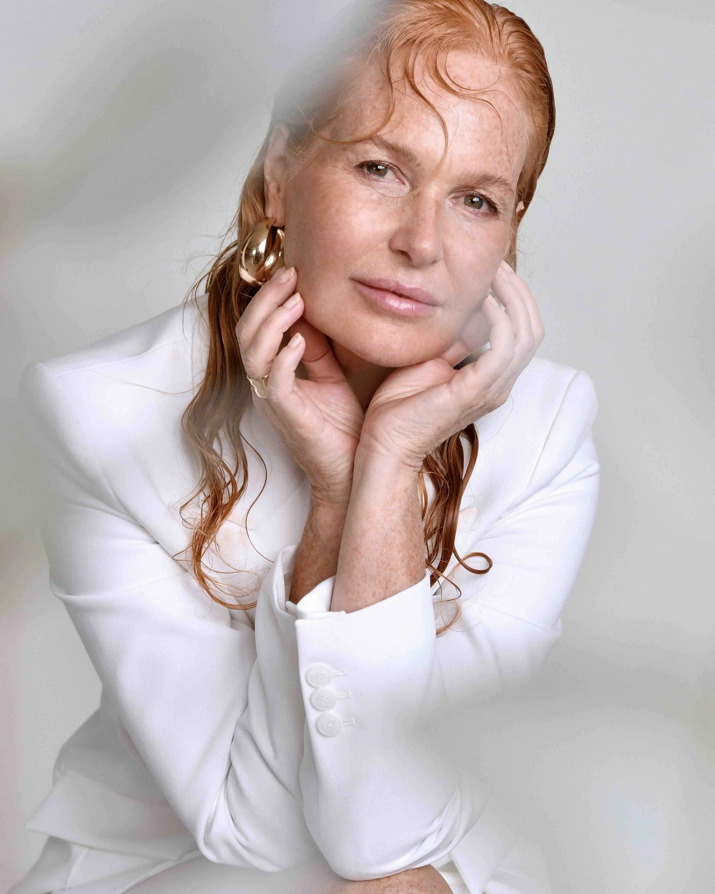 Eine Frau mit roten, gewellten Haaren und Sommersprossen trägt eine weiße Jacke und große goldene Ohrringe, sie posiert vor einem neutralen Hintergrund, ihre Hände sind an ihrem Gesicht und sie schaut direkt in die Kamera.