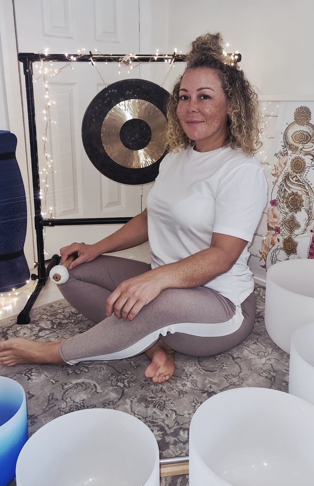A woman with curly hair and freckles sitting on a patterned rug, holding a mallet, with crystal singing bowls and a gong behind her, decorated with fairy lights and a flower-patterned backdrop.