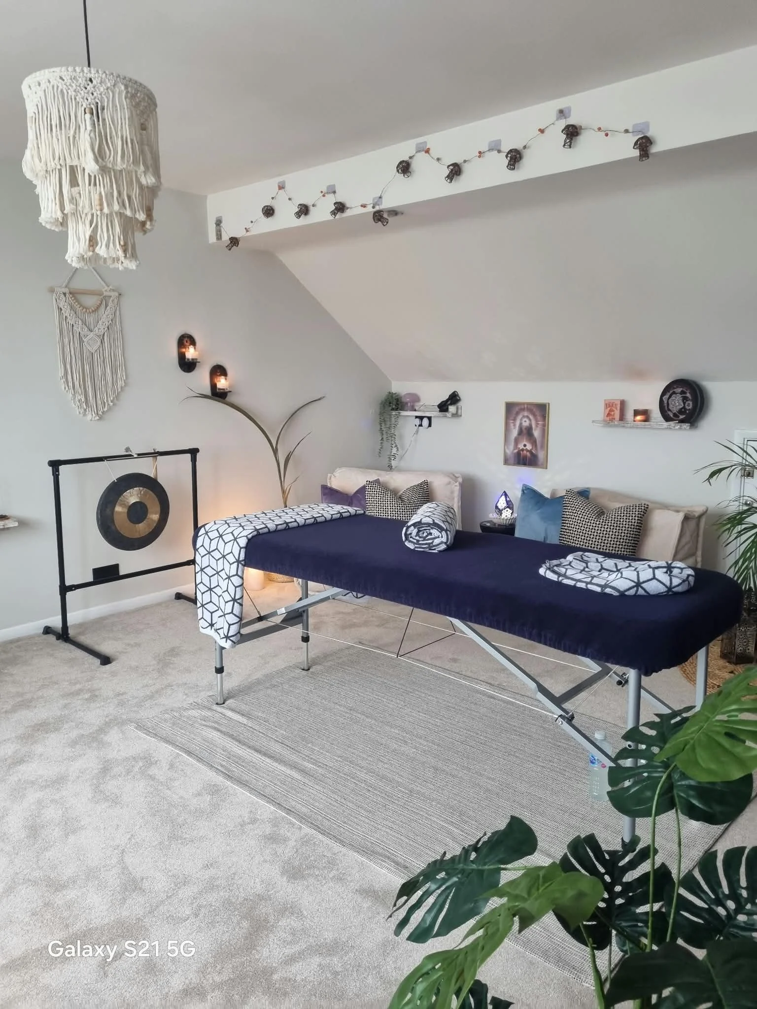 A massage room with a massage table covered in a dark blue sheet, surrounded by plants, artwork, and decorative lighting, with a hanging macrame ornament and string lights on the wall.