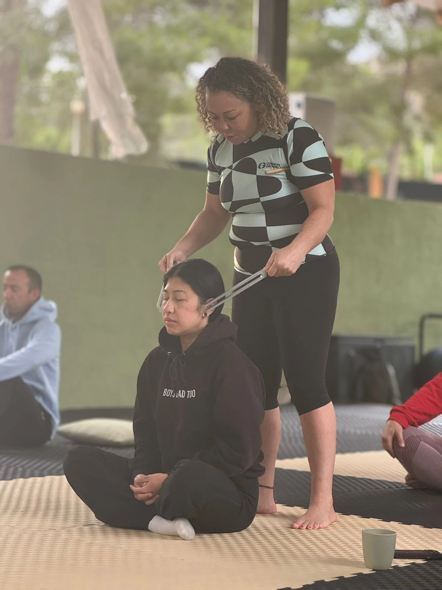 A woman performing a seated meditation or yoga pose while an instructor presses her head with a lever in a group class. The woman is sitting cross-legged with her eyes closed, wearing a black hoodie. The instructor stands behind her, using a device to gently press on her head while she maintains the pose. Other participants sit nearby in a room with large windows, natural light, and trees outside.